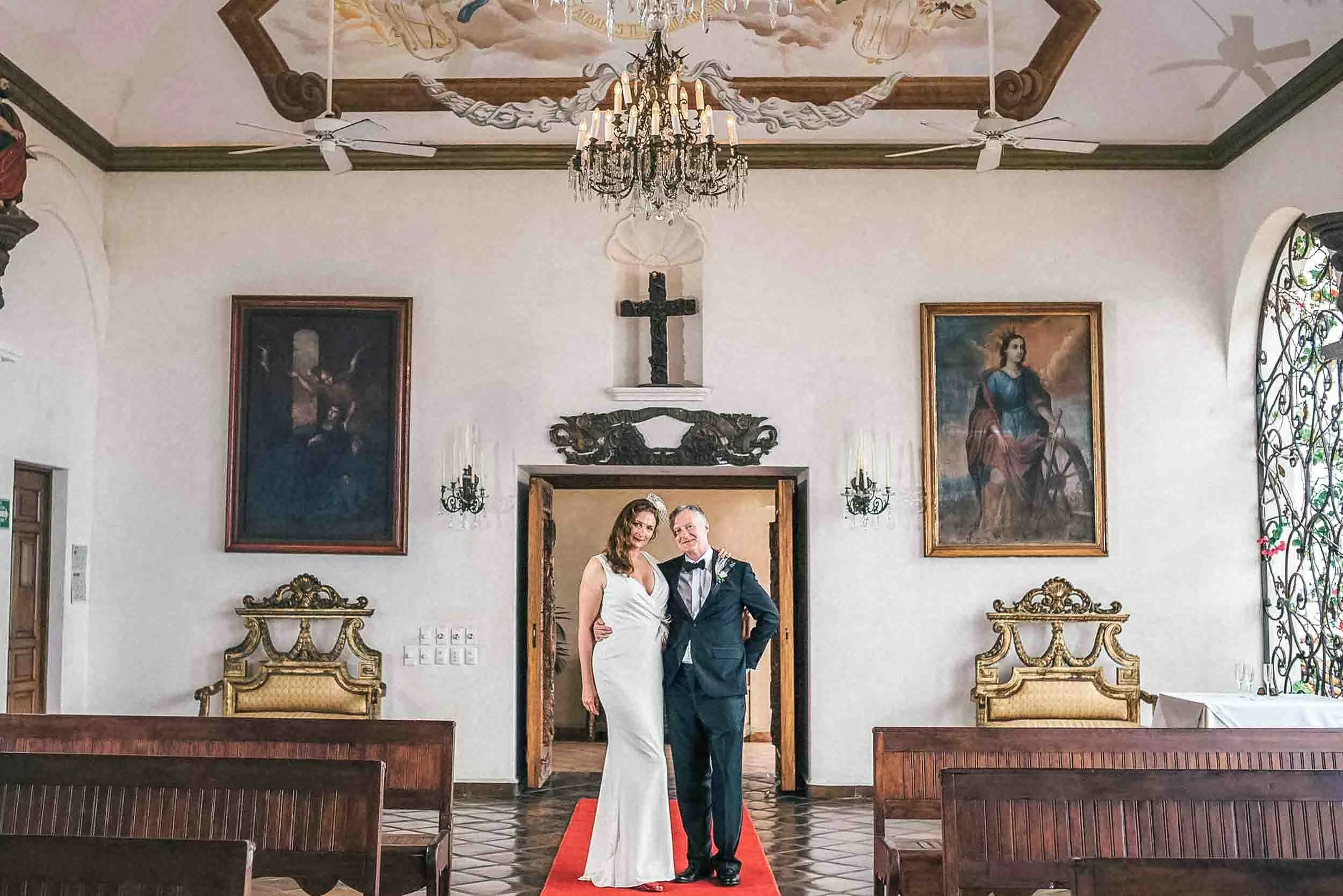 A bride and groom standing on a red carpet inside La Capella, smiling at the camera. The venue has ornate gold chairs, large paintings on the walls, a chandelier hanging from the ceiling, and a cross on the wall behind them.