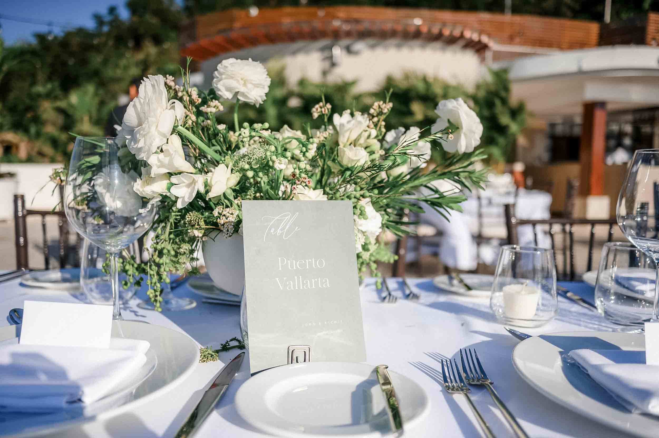 An outdoor wedding reception table decorated with a floral centerpiece of white flowers and greenery, candle, wine glasses, and place settings. A card with the text 'Table Puerto Vallarta' is visible.