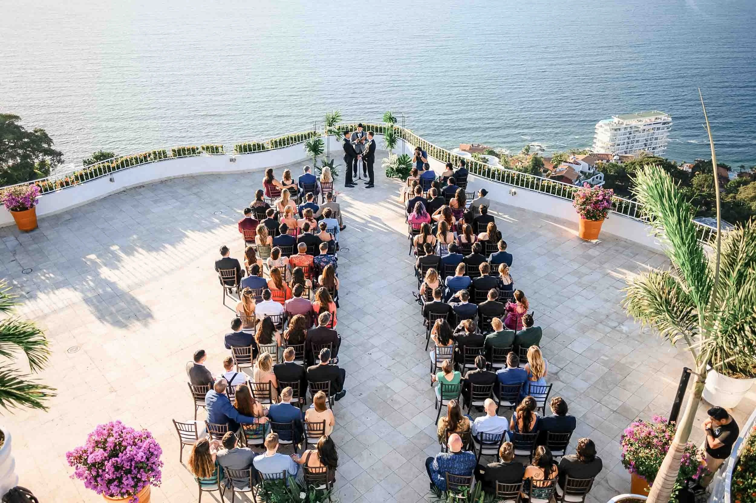 A destination wedding ceremony taking place outdoors on a terrace overlooking the ocean, with guests seated facing a couple of groomsmen at the Grand Miramar resort.