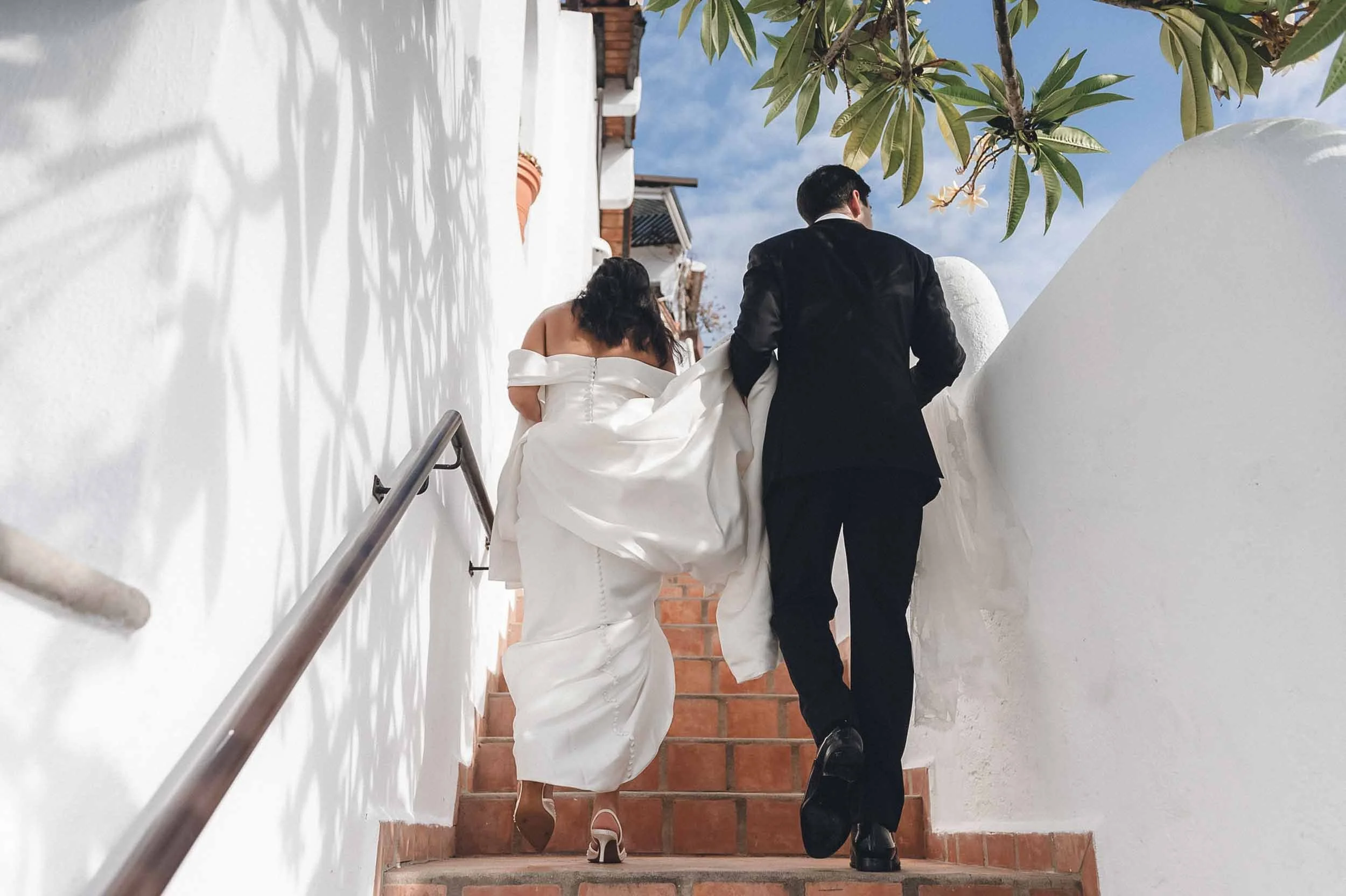 A bride and groom walking up a staircase outdoors with white walls and brick steps. The bride is wearing a strapless white wedding gown, and the groom is dressed in a black tuxedo. The scene is shaded by tree leaves and a blue sky is visible.