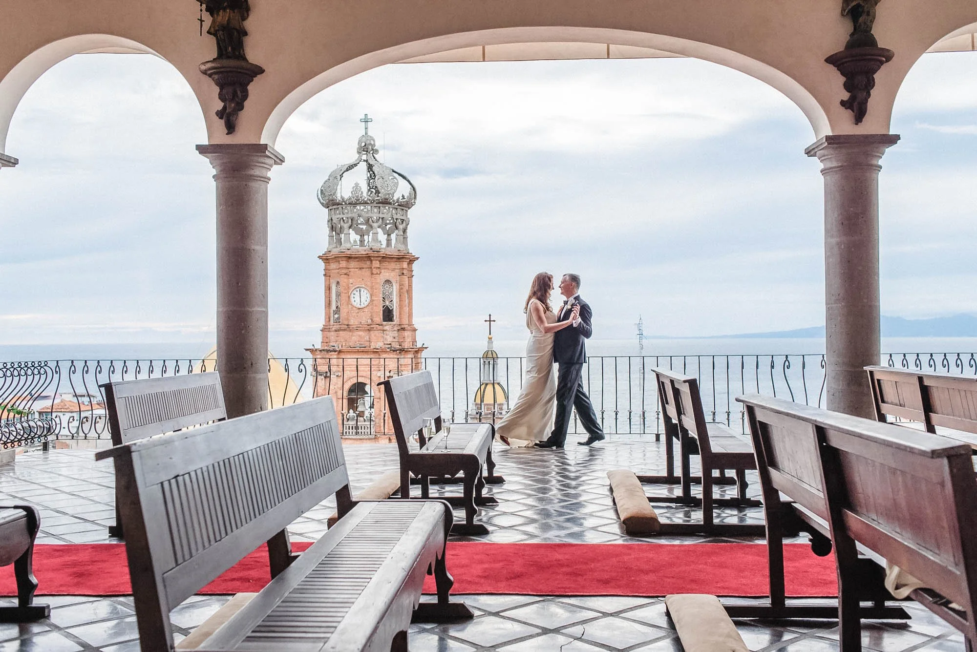 Bride and groom dancing on a balcony with a view of a Puerto Vallarta church with the clock tower and the sea in the background, at La Capella venue in Puerto Vallarta.