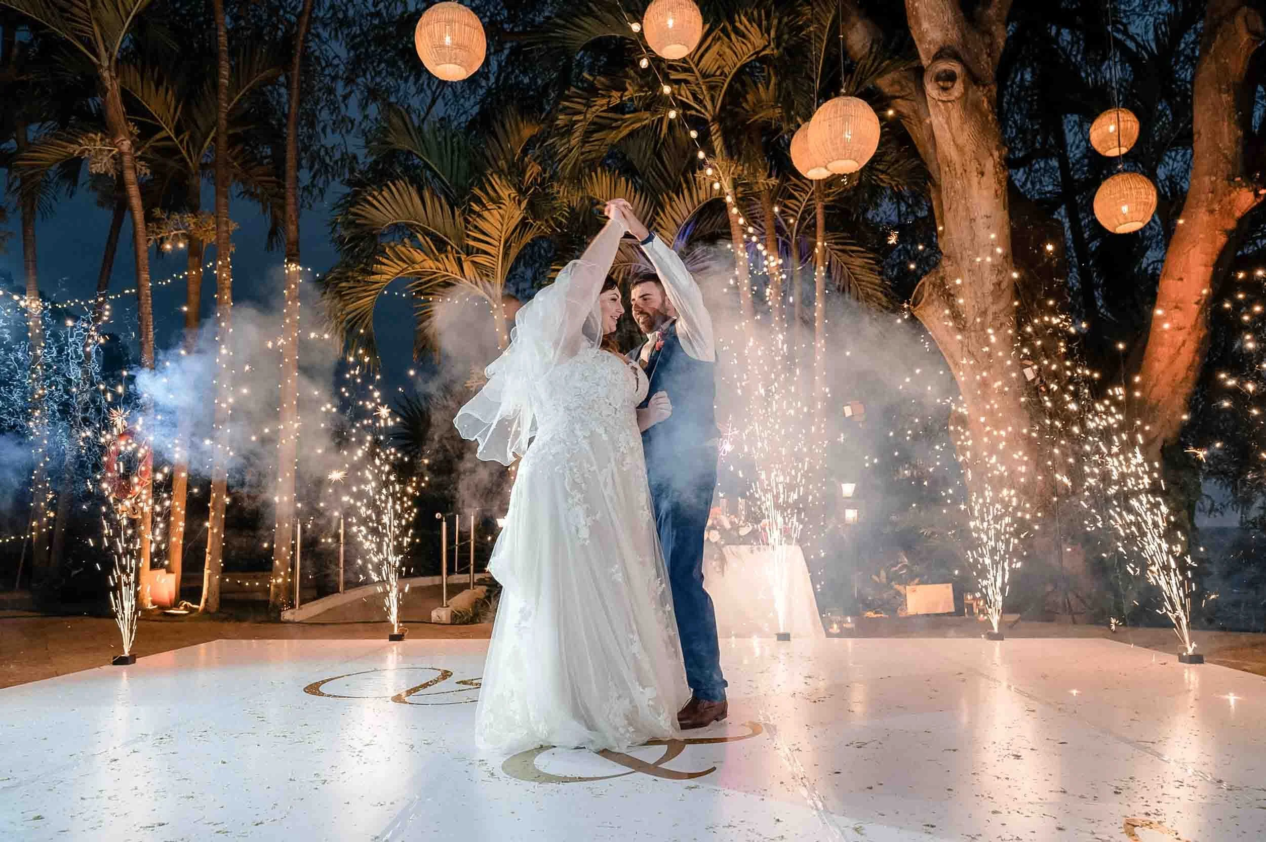 A bride and groom dancing at their wedding reception during nighttime, surrounded by sparklers and string lights with trees and hanging lanterns in the background.