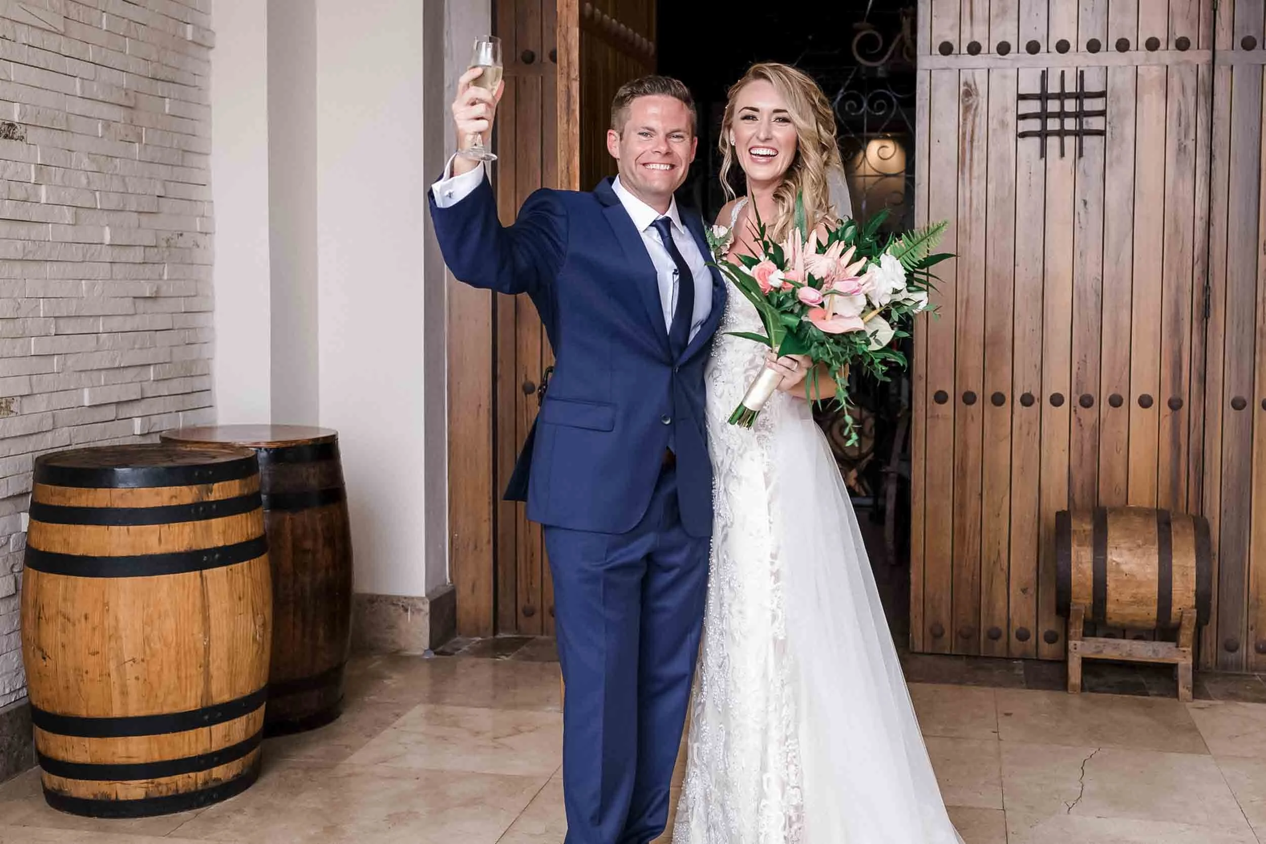 A newlywed couple celebrating outside, the groom in a blue suit holding a glass of champagne, the bride in a wedding gown holding a bouquet of pink and white flowers, both smiling.