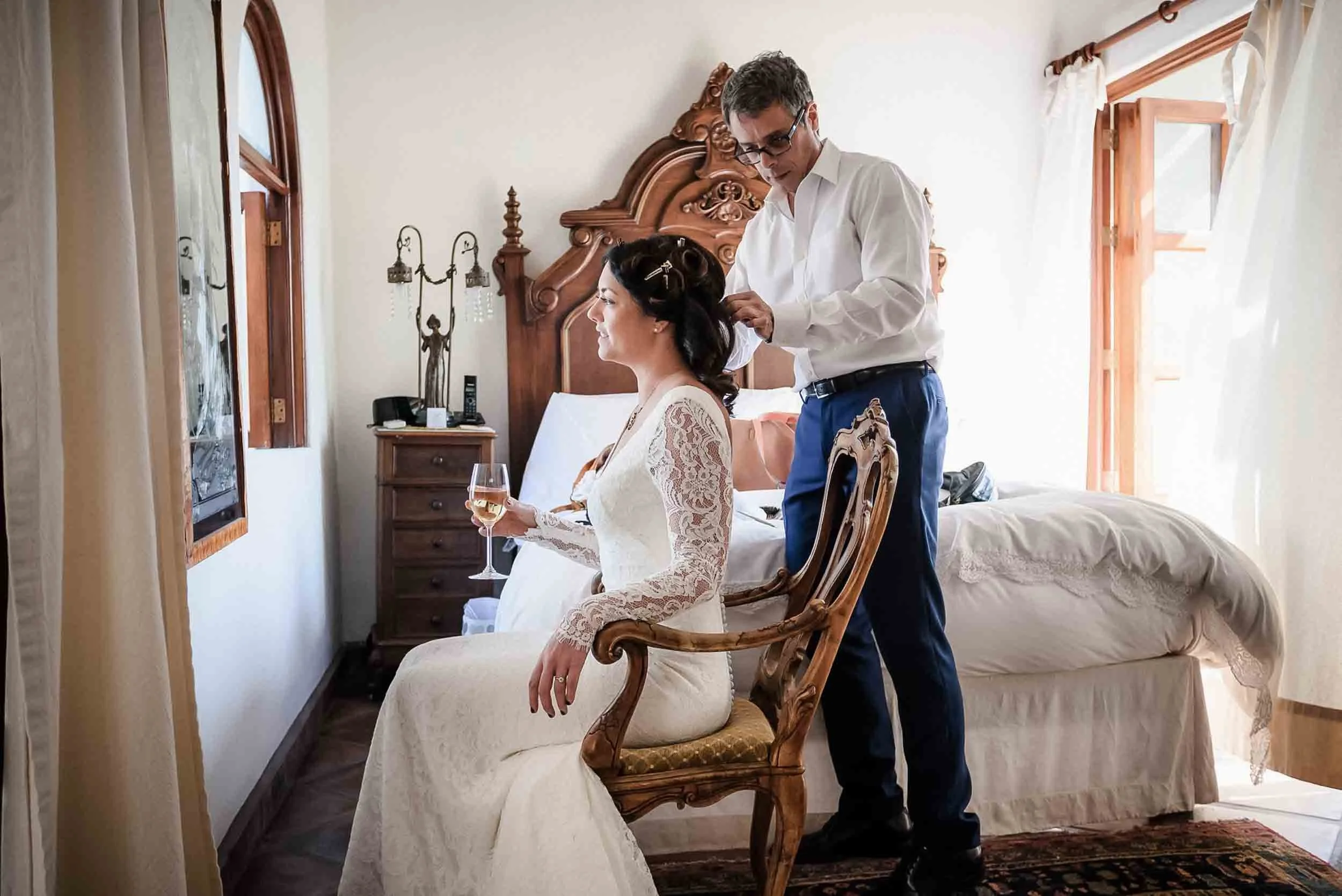A woman in a wedding dress sitting in a carved wooden chair, holding a glass of white wine, as a man stands behind her adjusting her hair in a bedroom with a large bed, wooden furniture, and windows with curtains.