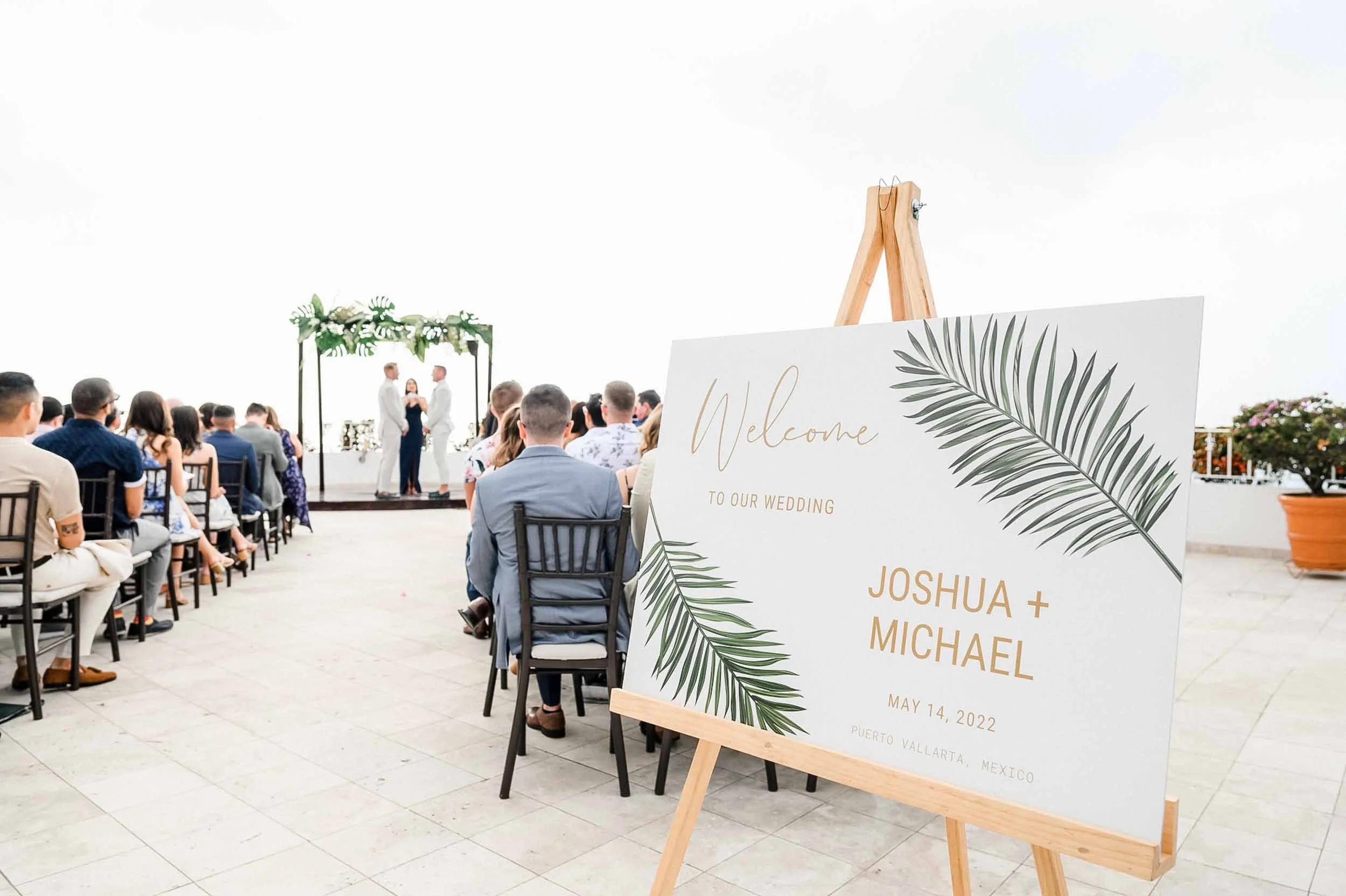 Gay wedding ceremony taking place on a terrace of the Grand Miramar resort.  Welcome sign in the foreground displays the names Joshua and Michael with the date May 14, 2022, and location Puerto Vallarta, Mexico