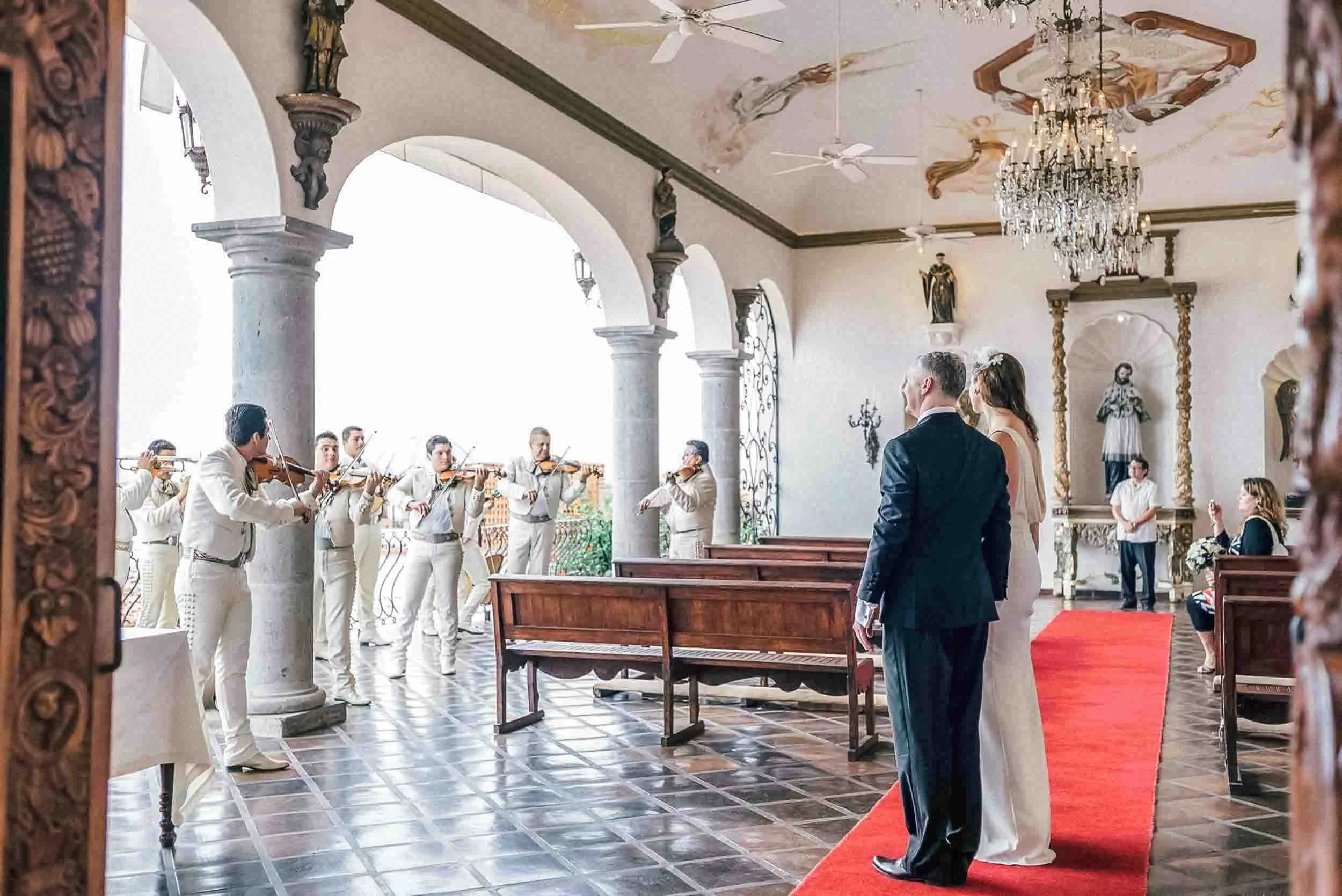 Bride and groom standing looking at the mariachis singing for them. The venue features large arched windows, a chandelier hanging from the ceiling, ceiling fans, and religious statues on the walls.