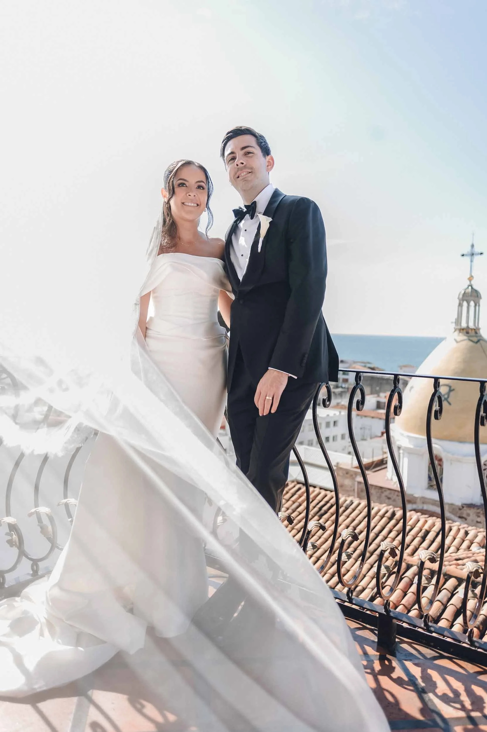 A bride and groom in wedding attire standing on a balcony and a church tower in the background.