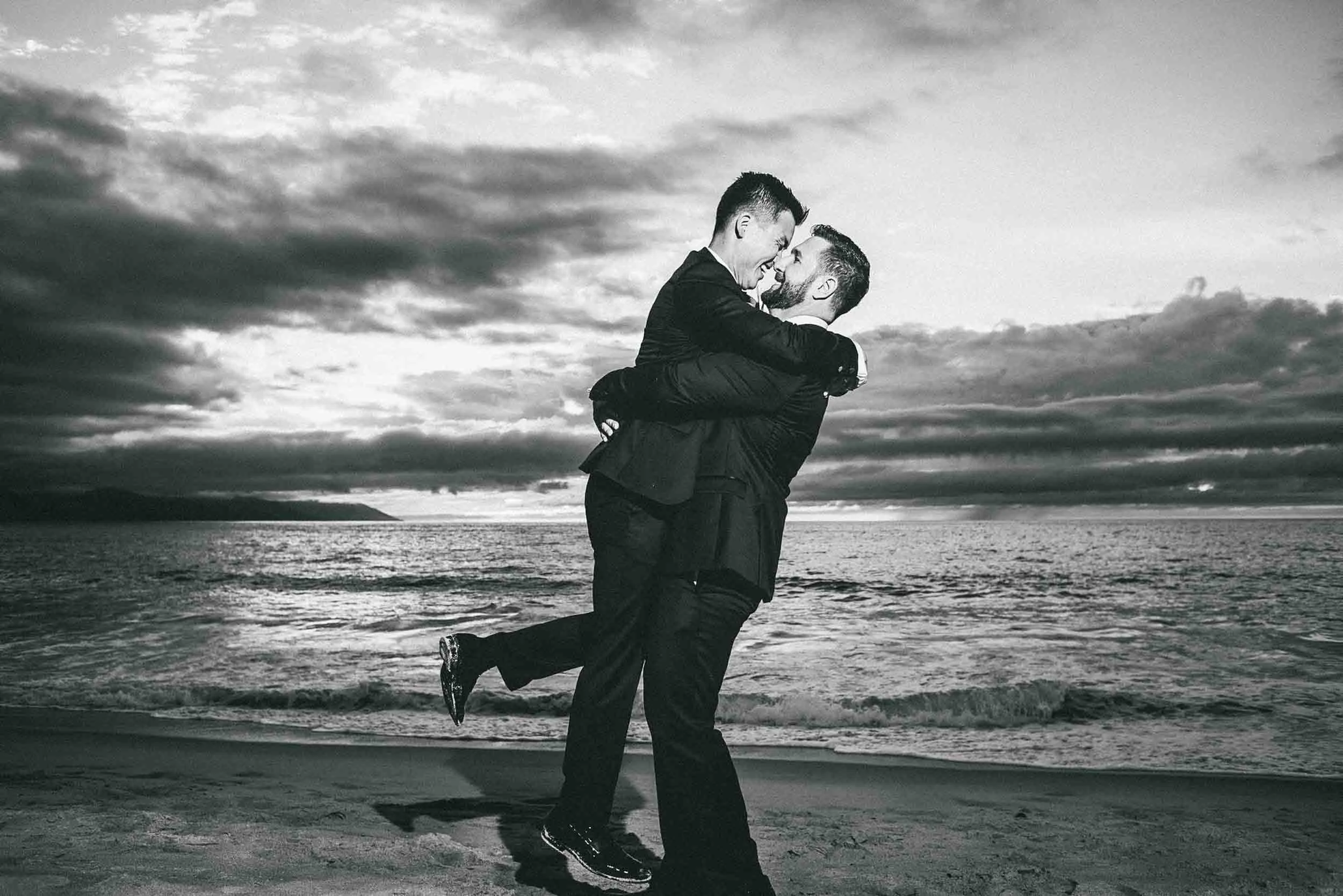 Two grooms with one lifting the other in a joyful embrace, with a cloudy sky and ocean in the background.