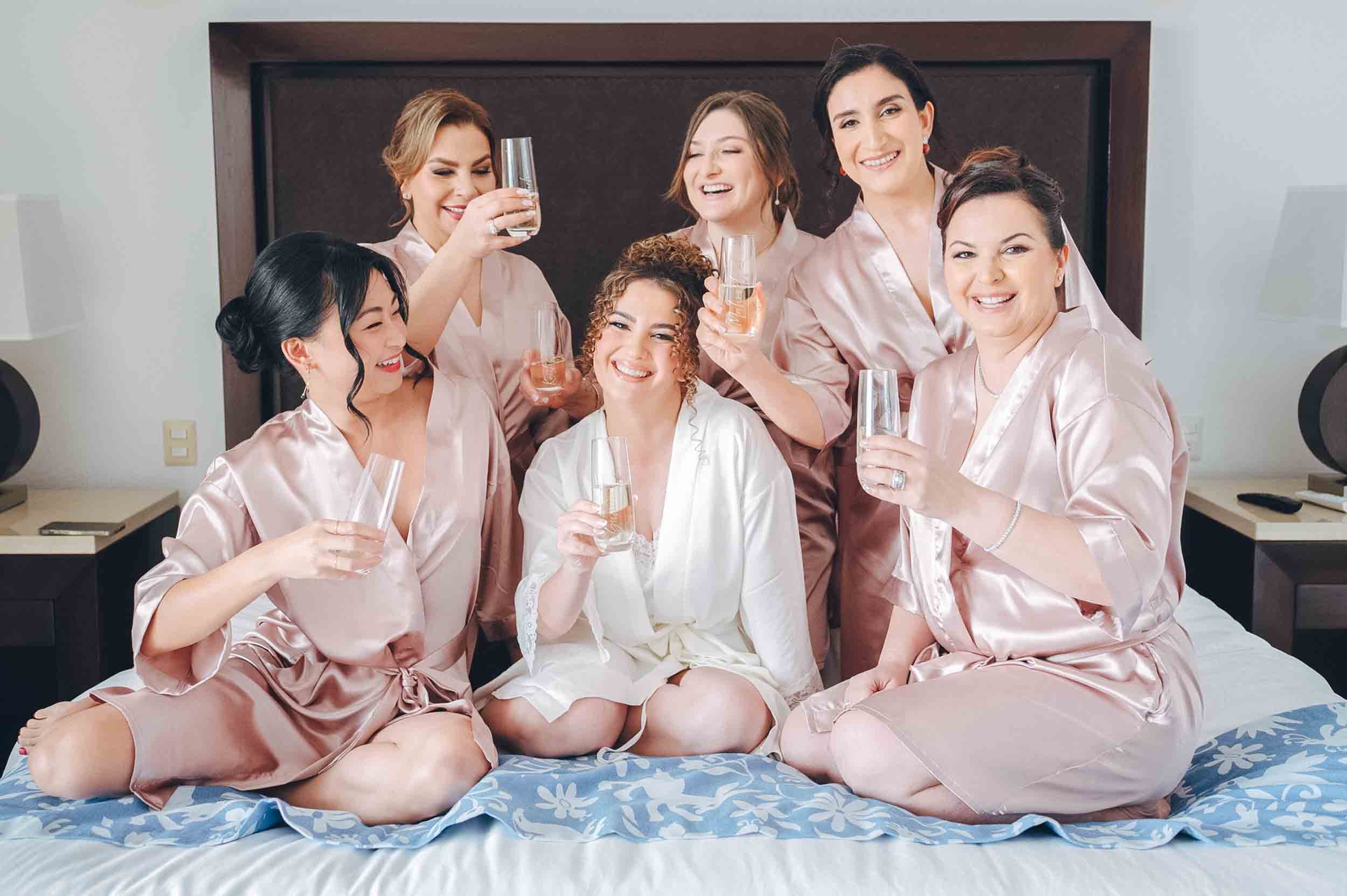 Group bridesmaids and bride in satin robes celebrating with glasses of champagne before getting ready for the wedding ceremony.
