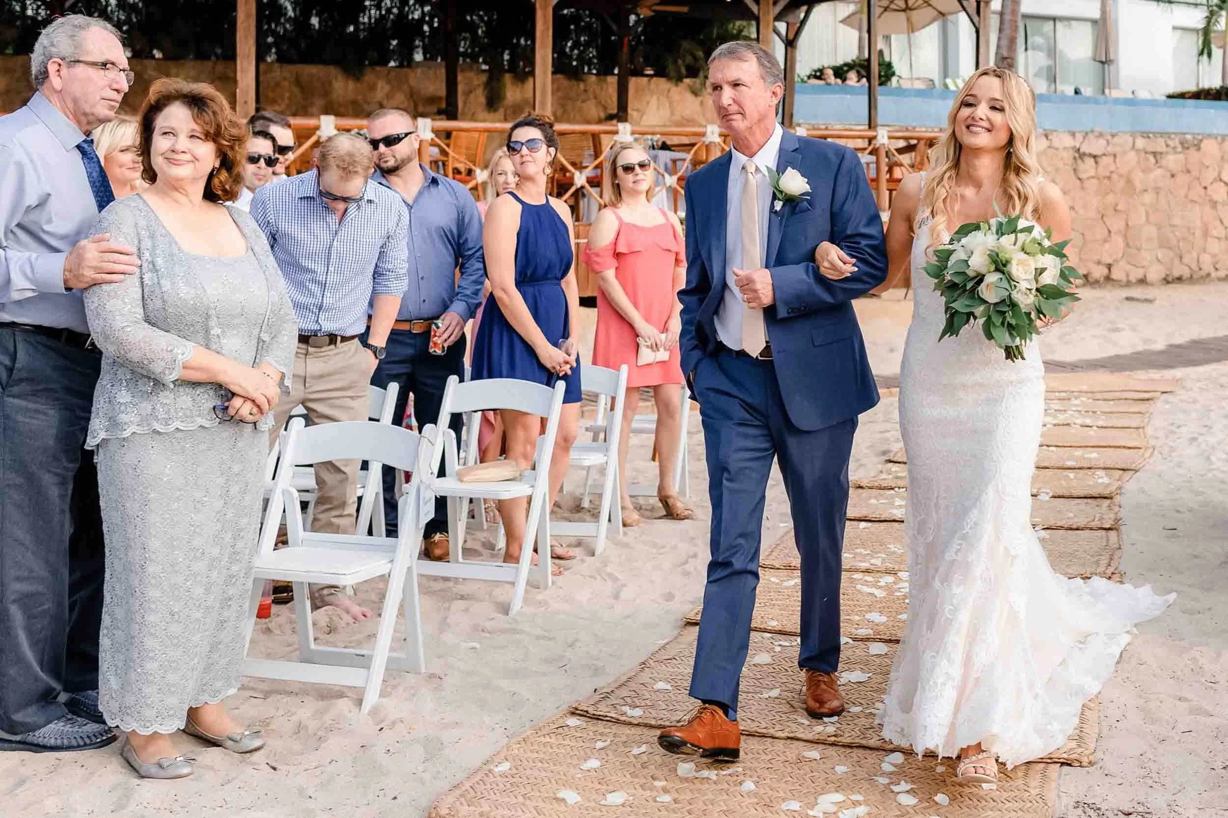 Bride holding a bouquet walks down the aisle, accompanied by her father during her destination wedding..