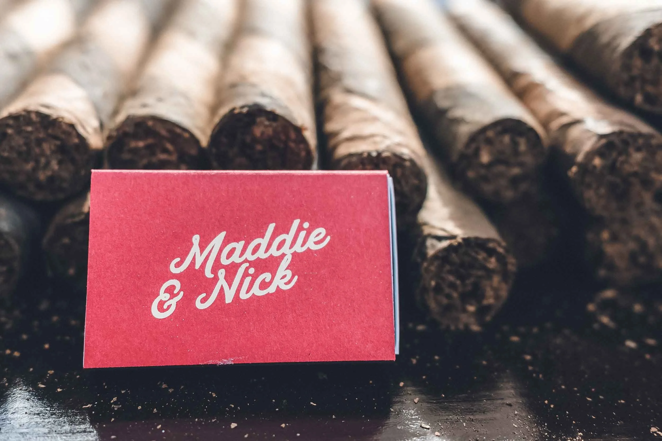 Close-up of a match box with white cursive text reading 'Made with & Nick,' in front of several hand rolled cigars as wedding keepsakes.