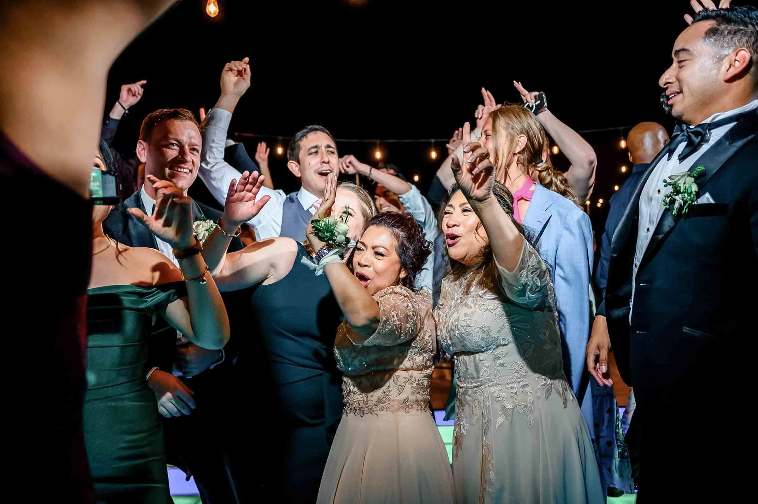 Group of well-dressed people celebrating and dancing during  a wedding party with string lights overhead.
