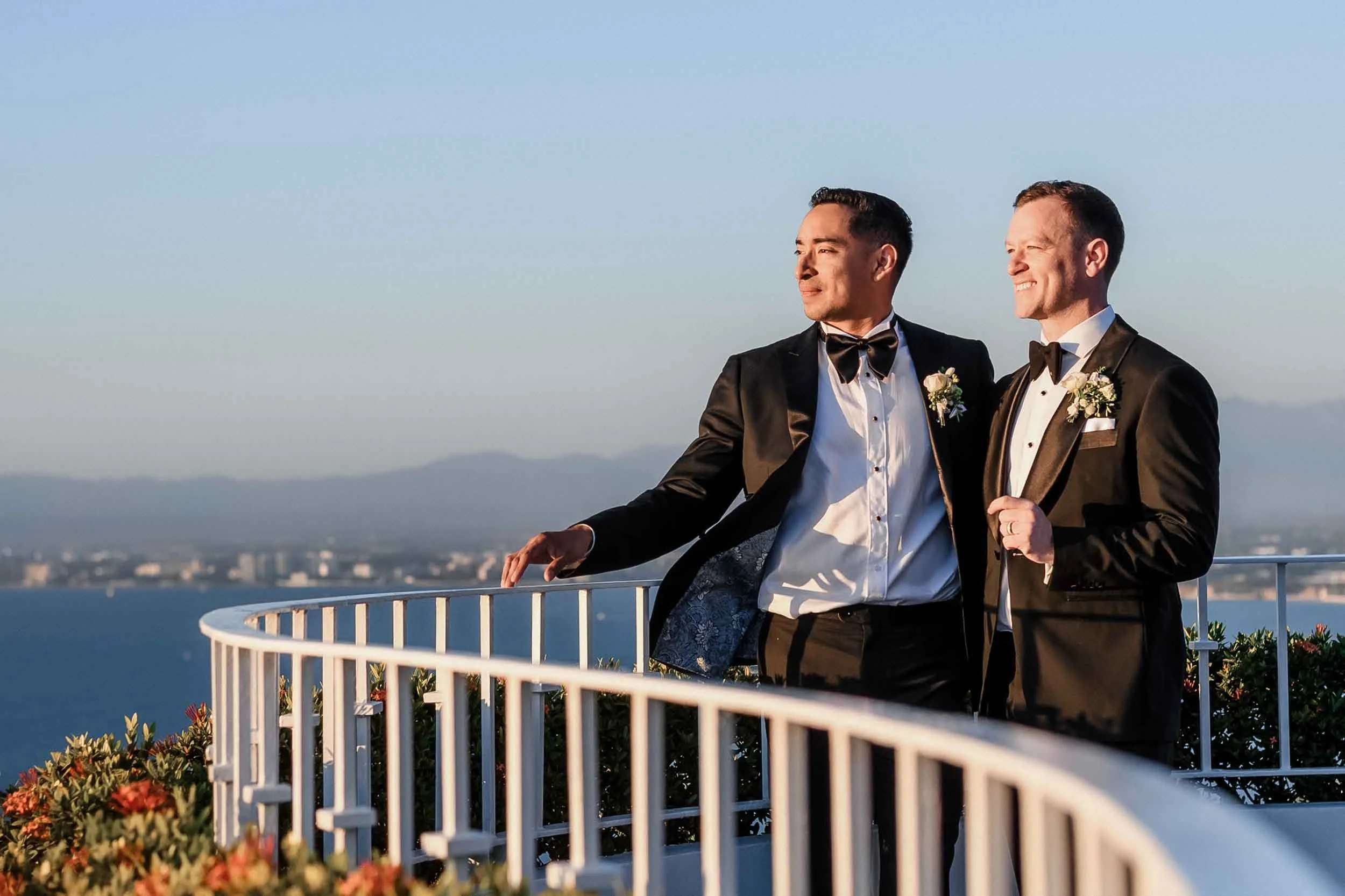 Grooms just married standing on a balcony overlooking the ocean with mountains in the background, after their wedding ceremony.