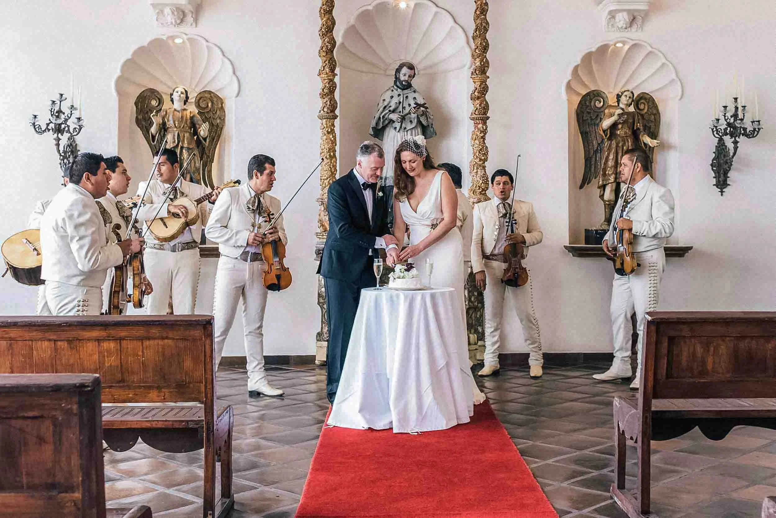 Bride and groom cutting their wedding cake, on the sides are members of the mariachi singing to them. 