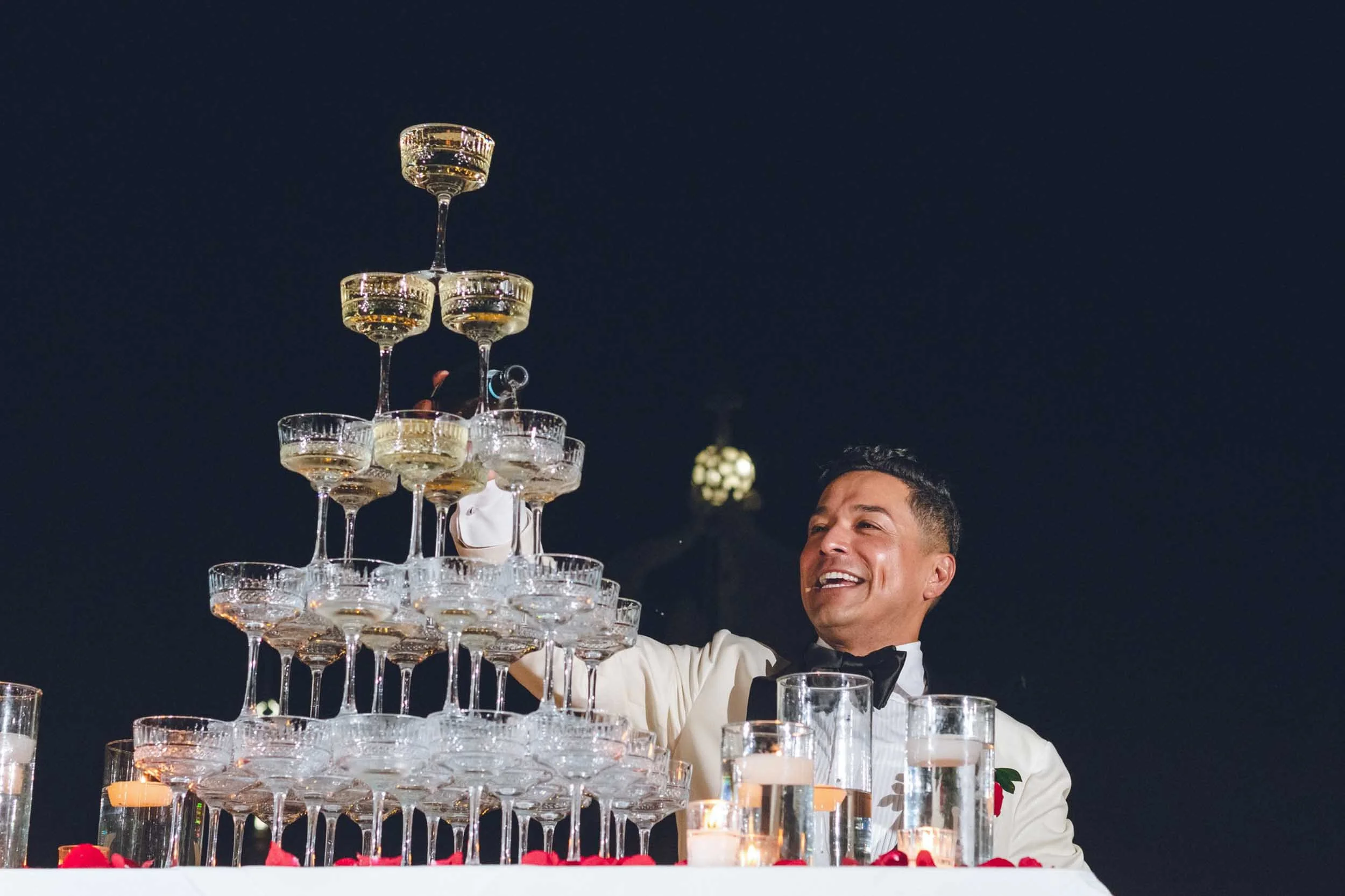 Groom is smiling while stacking champagne glasses on a table decorated with candles and rose petals during his wedding celebration.