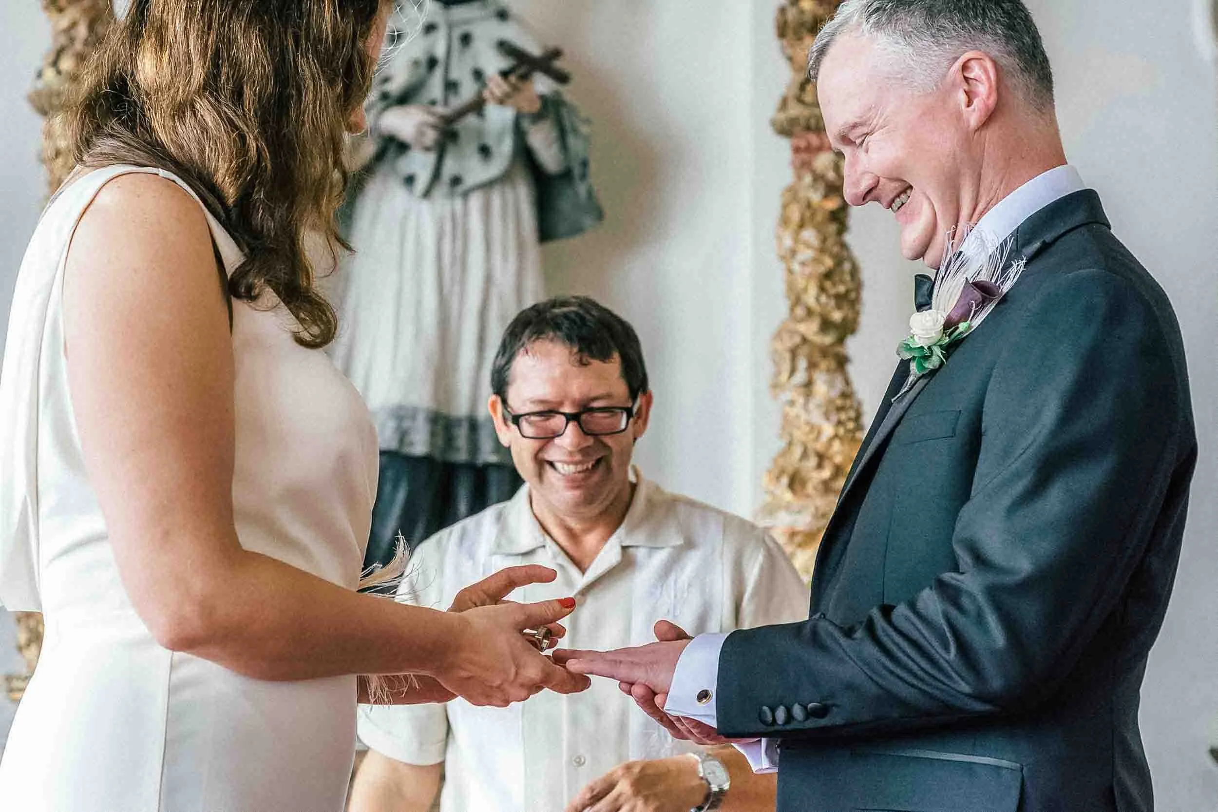 A wedding ceremony with a bride and groom exchanging rings, an officiant smiling, and a woman taking photos in the background.