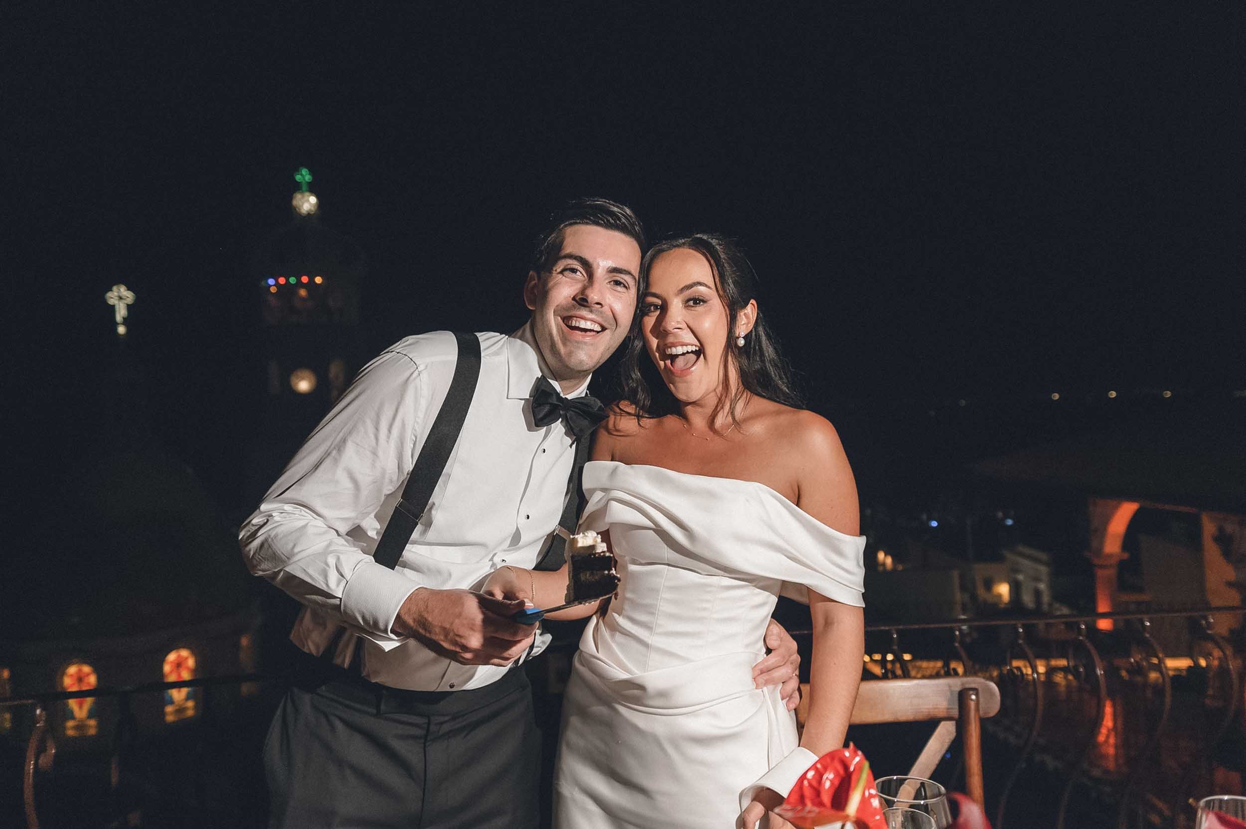 A happy couple at night, dressed in formal wedding attire, smiling for a photo on a rooftop or outdoor venue with city lights in the background.