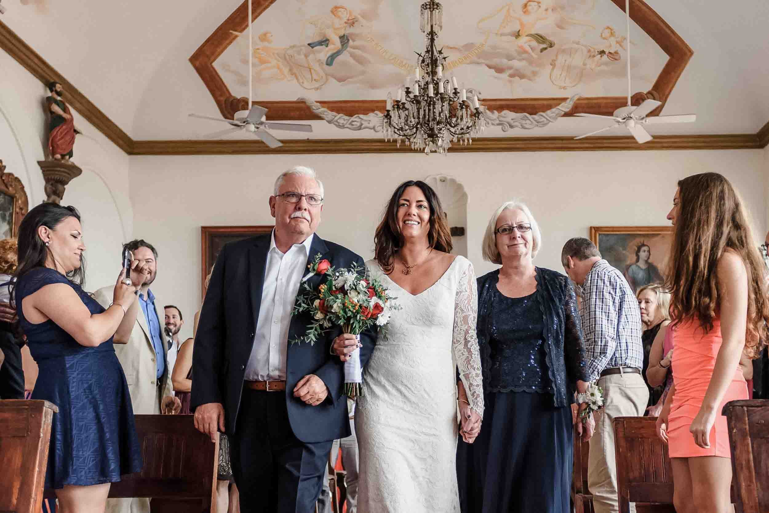 Parents walking a bride down the aisle surrounded by guests, in La Capella venue decorated with a chandelier and ceiling mural 