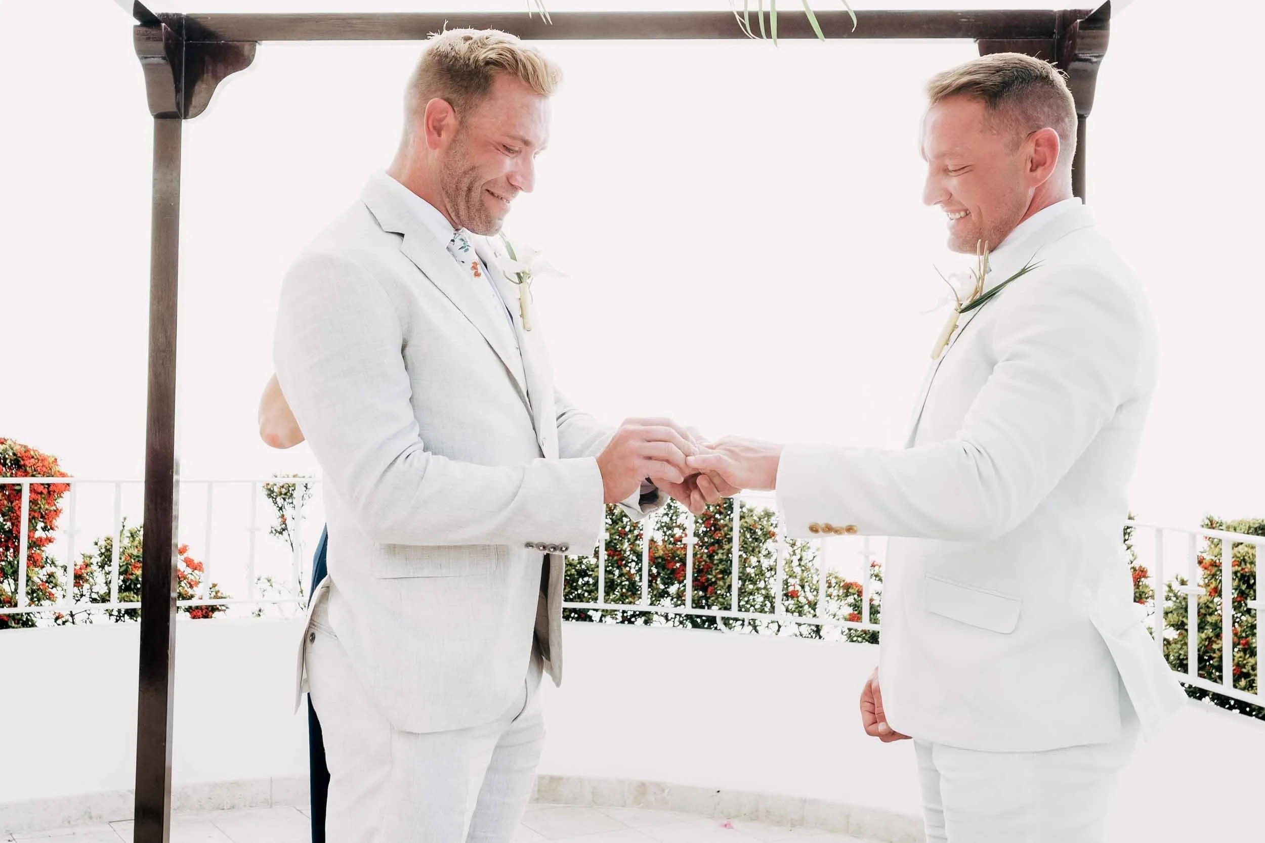 Grooms  exchanging rings during their wedding ceremony outdoors, with a wooden arch and potted flowers in the background.