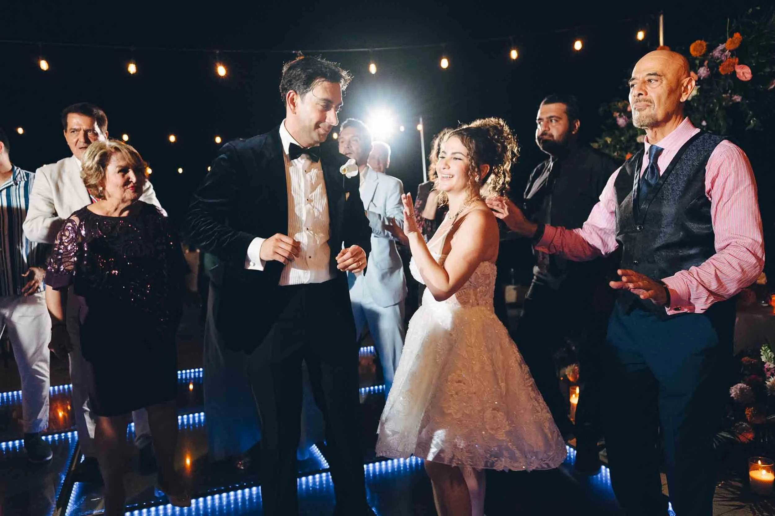 A wedding reception at night with string lights, showing the bride and groom dancing and smiling at each other, surrounded by guests.