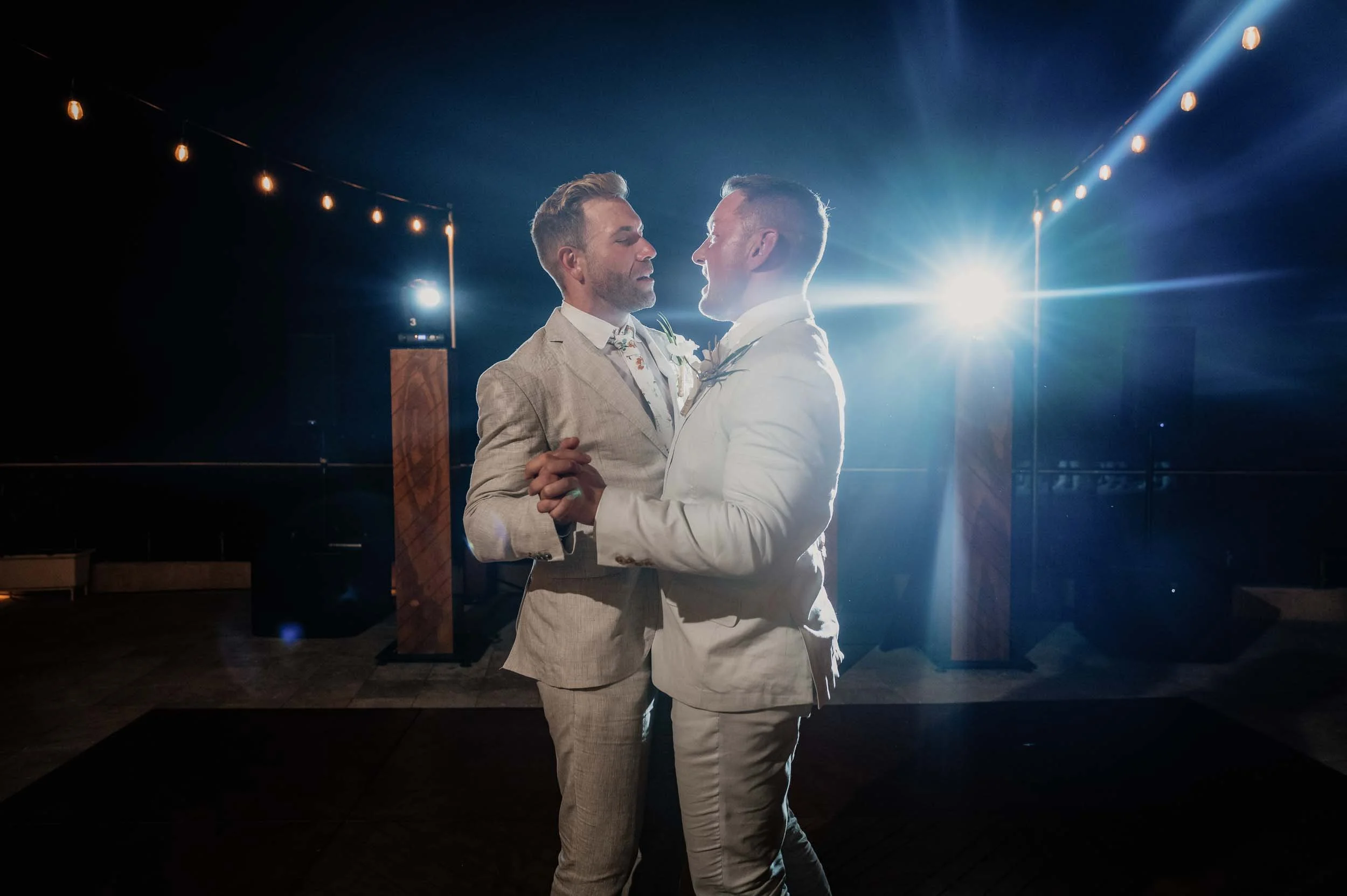 Grooms on dance floor during their first dance at their wedding reception, with bright lights shining behind them.
