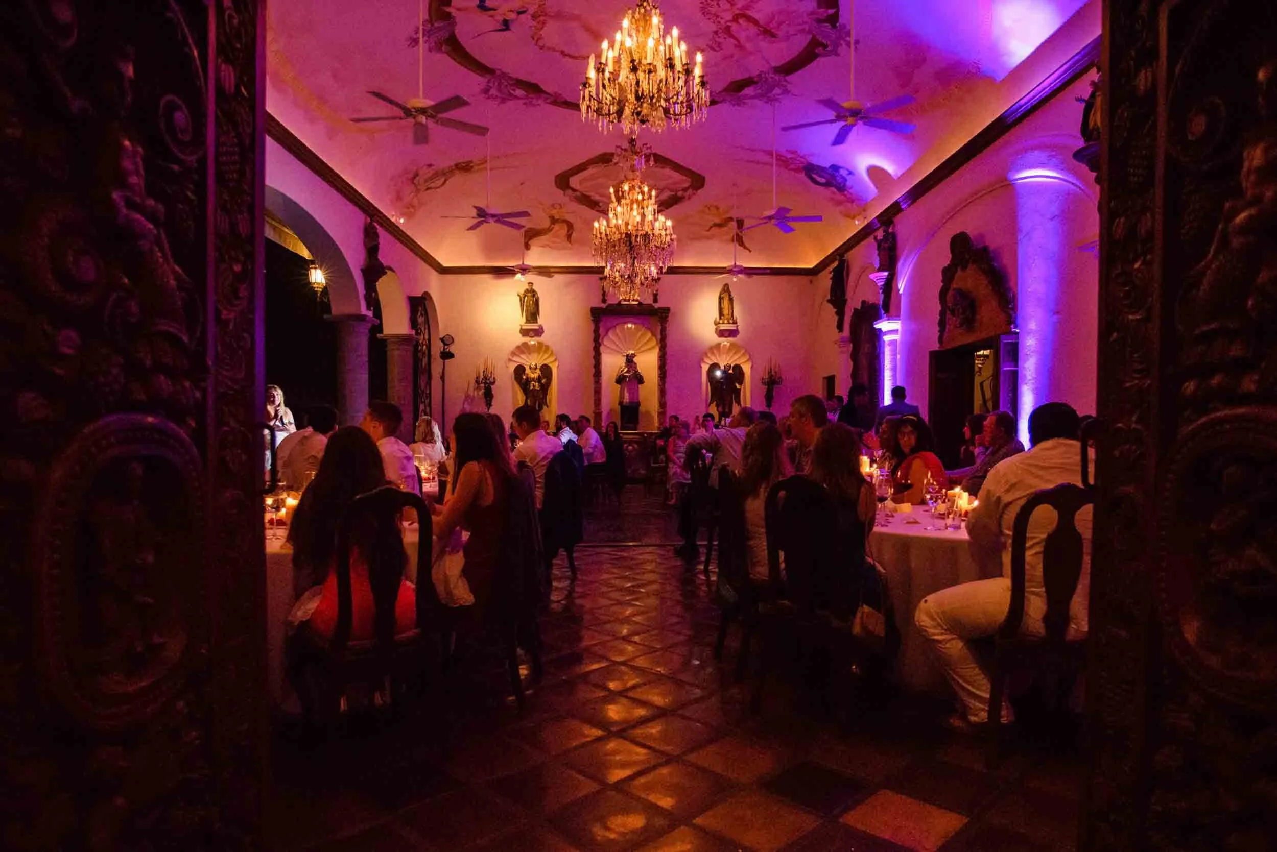 People dining during wedding reception in a luxurious, ornately decorated room with chandeliers and classical statues, illuminated by warm and purple lighting.