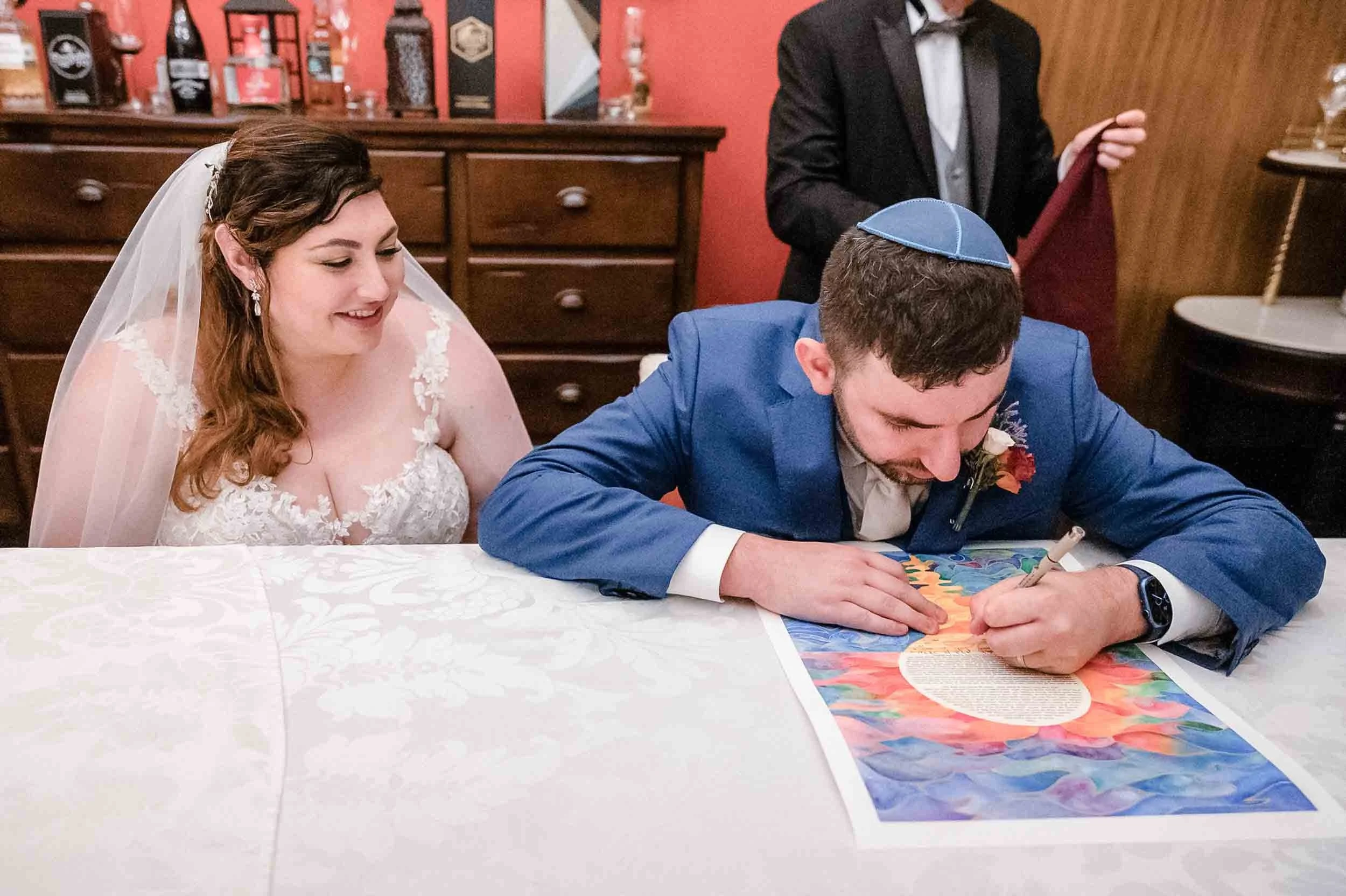 A bride and groom at their wedding,  smiling as they sign the Ketubah. The groom is signing the document while wearing a blue suit and a kippah, with a colorful picture or painting on the table.