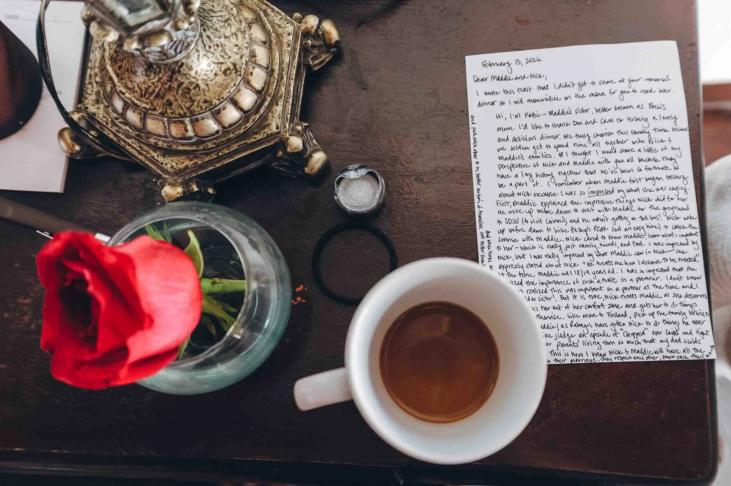 A wooden table holding a red rose in a small glass vase, a handwritten letter, a white coffee cup filled with coffee, and two small glasses of tea. 