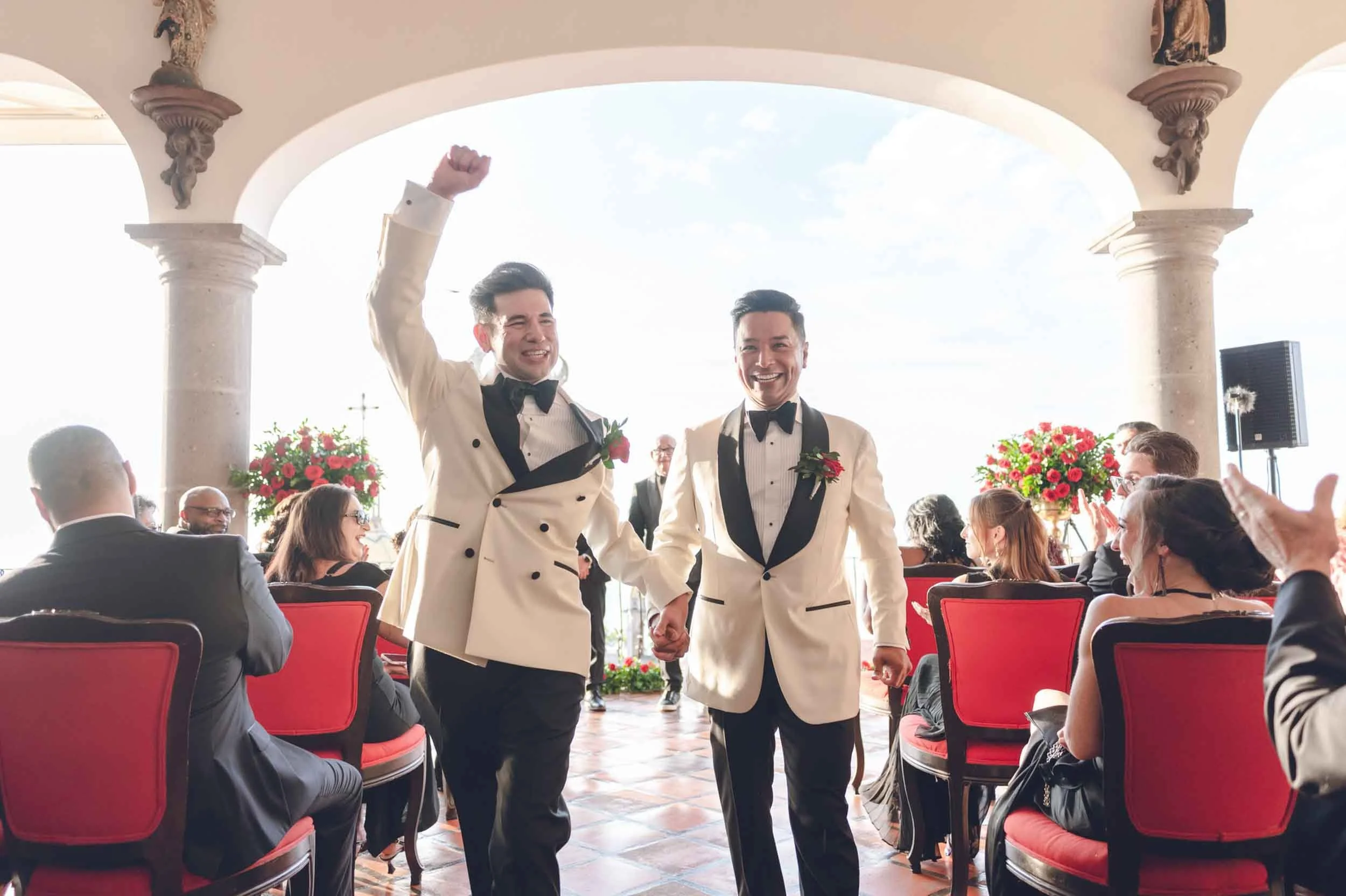 Gay couple in tuxedos walking hand in hand exiting their wedding ceremony in a sunny indoor venue, with guests seated around them and floral arrangements in the background.