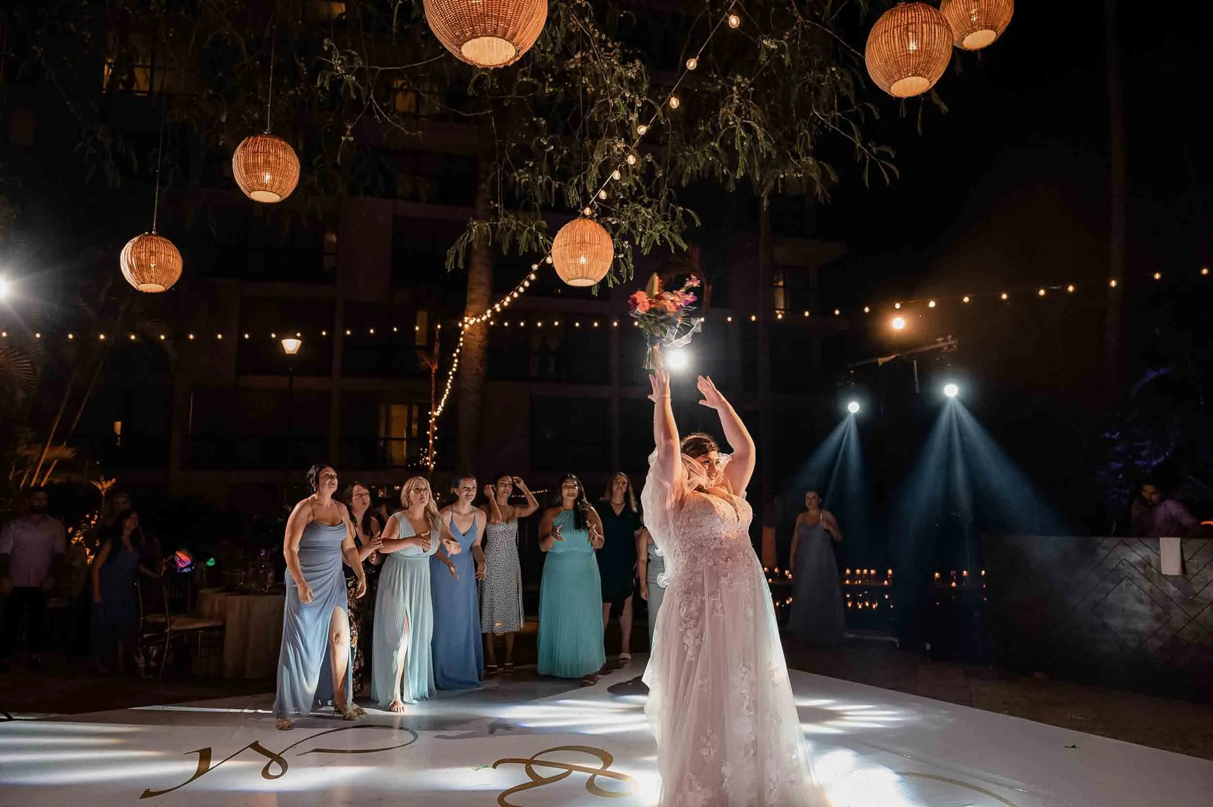Bride in a white lace wedding dress tossing a bouquet to the guests at an outdoor nighttime wedding reception with hanging lanterns and string lights at the Hyatt Ziva Puerto Vallarta.