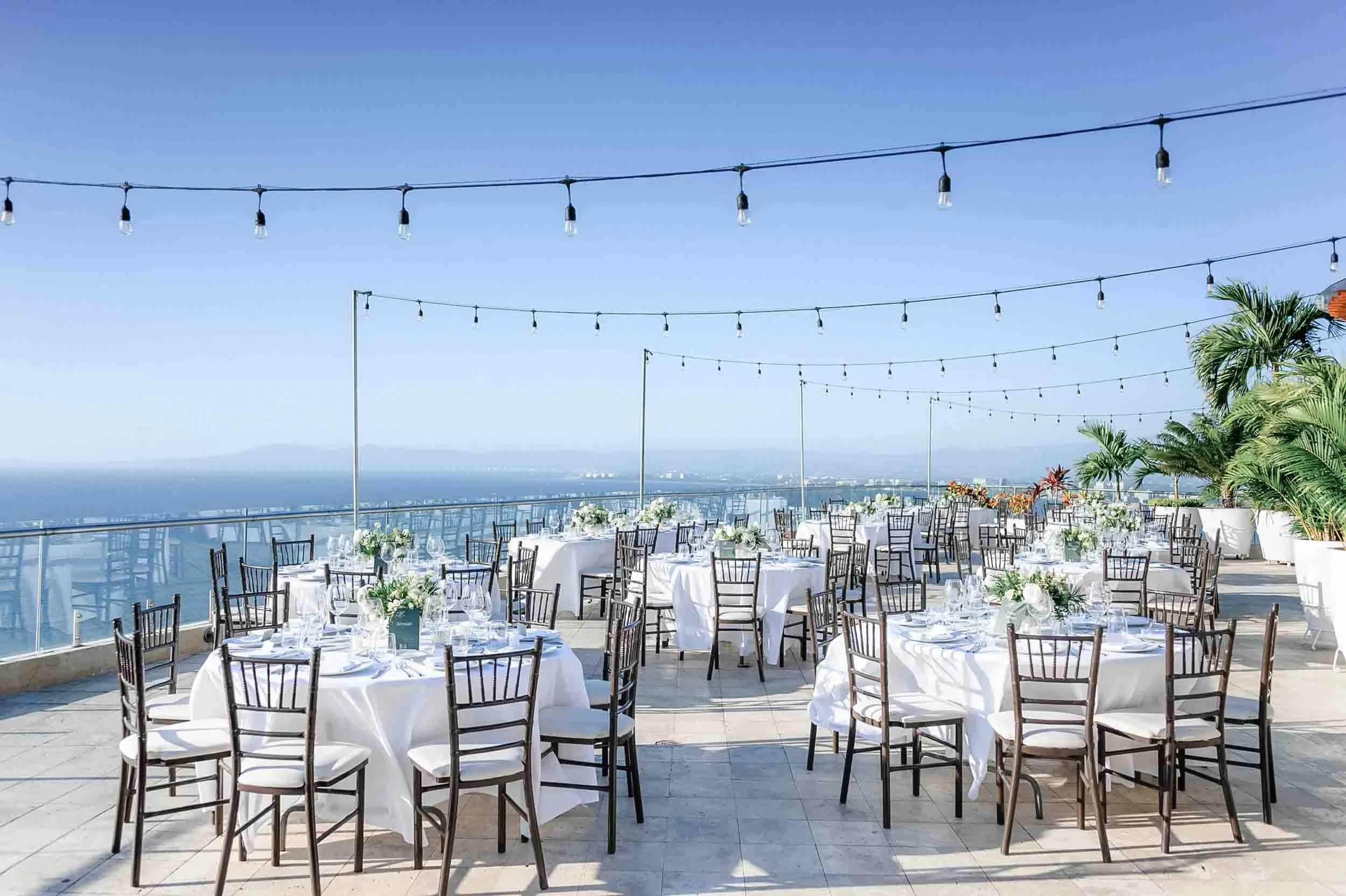 Outdoor wedding banquet setup with round tables covered in white tablecloths, black chairs, and floral centerpieces, overlooking a scenic ocean view with clear blue sky and string lights overhead.
