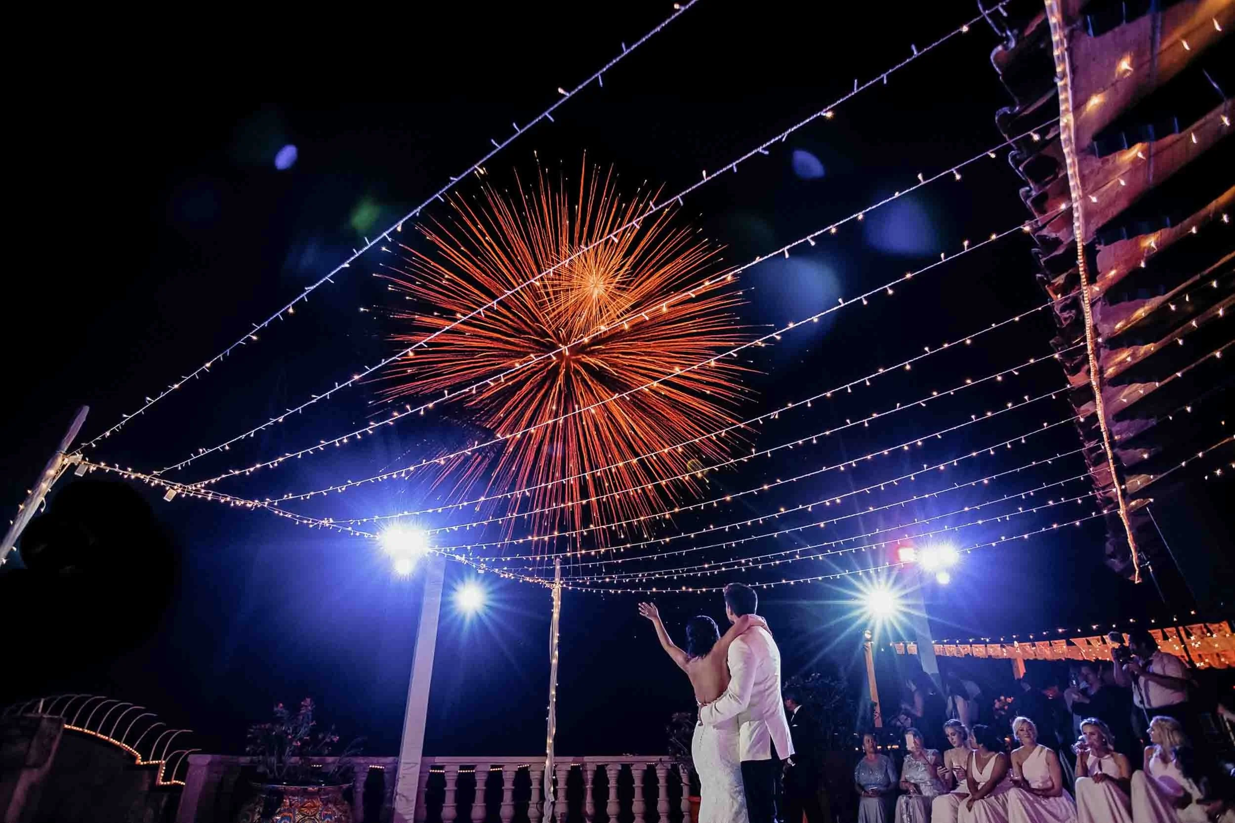 Night scene at a wedding with fireworks in the sky, string lights overhead, and the bride and groom.