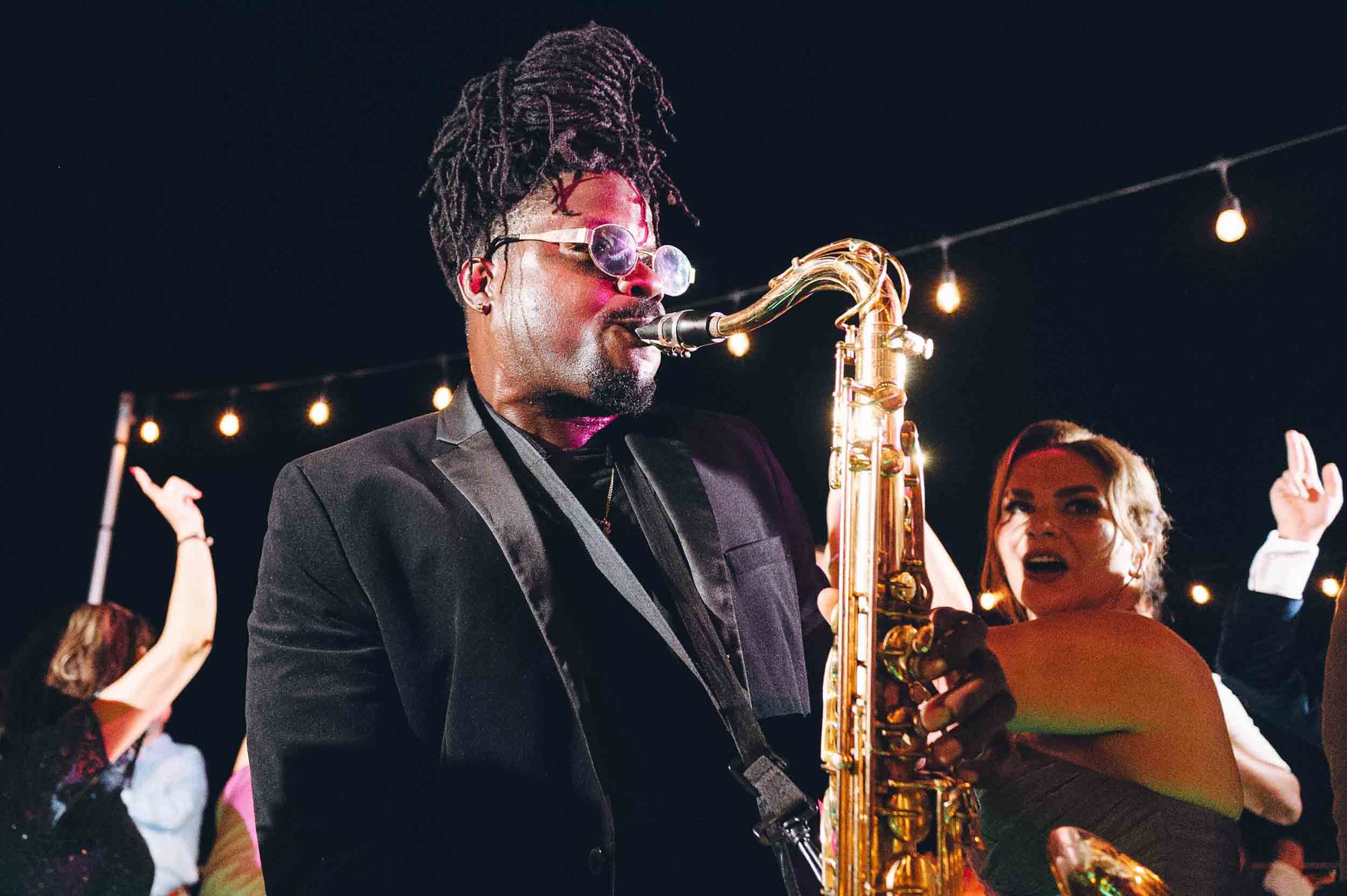 A man with dreadlocks, wearing sunglasses and a suit, playing a gold saxophone at night during a wedding reception.