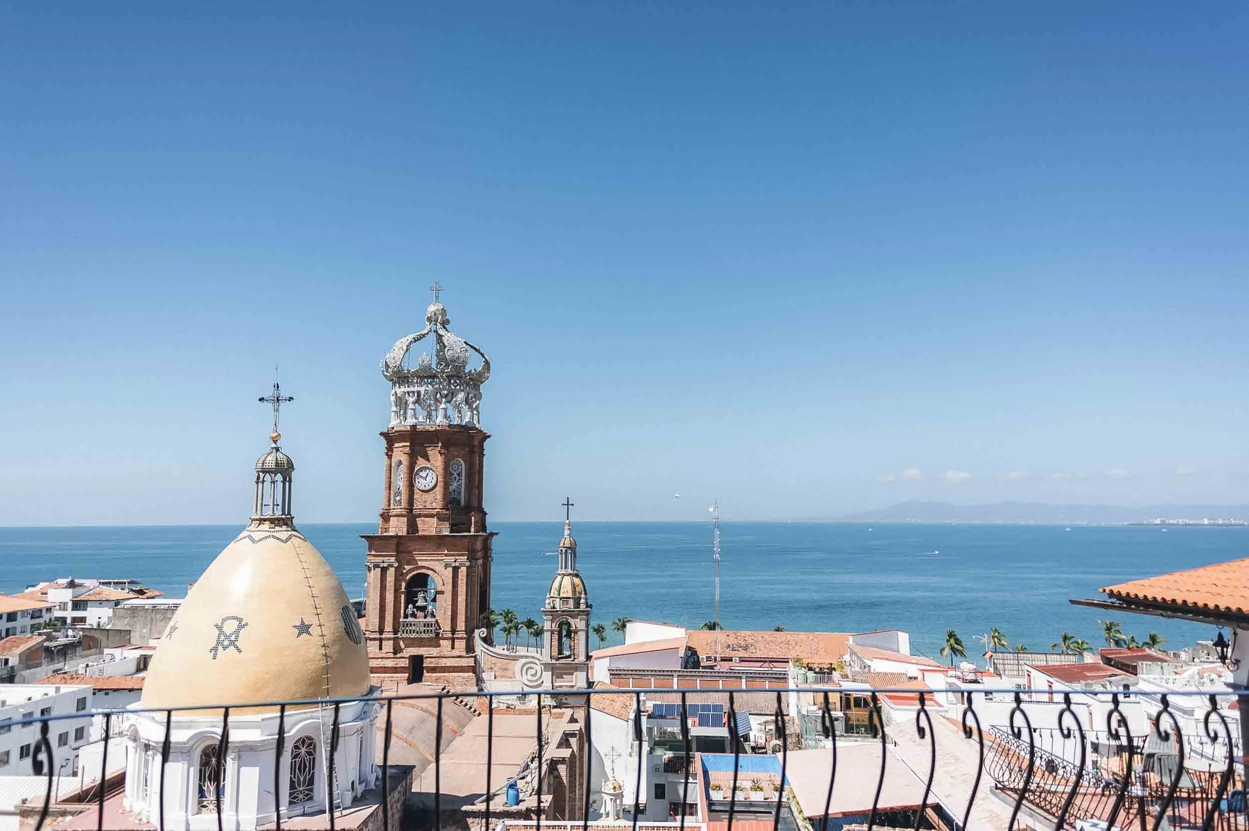 Church steeples and domes overlooking the ocean, with a blue sky above and a balcony railing in the foreground in Puerto Vallarta Mexico.