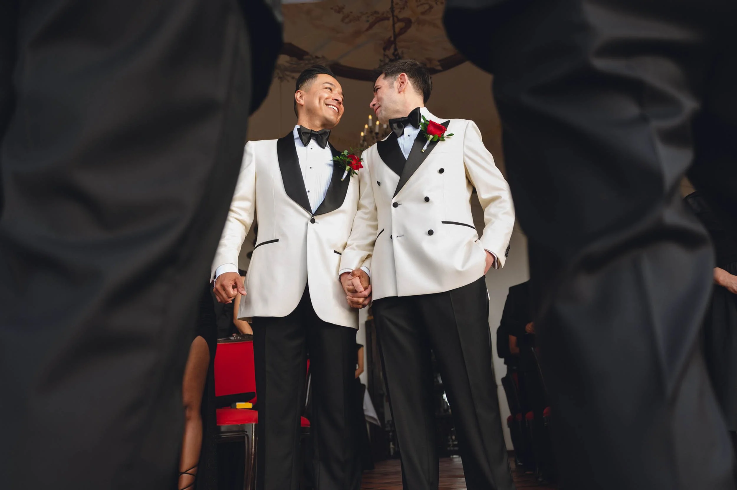 Gay newlyweds  in white tuxedos with black lapels and bow ties holding hands and smiling at each other during their wedding ceremony, seen from below through the legs of the officiant.