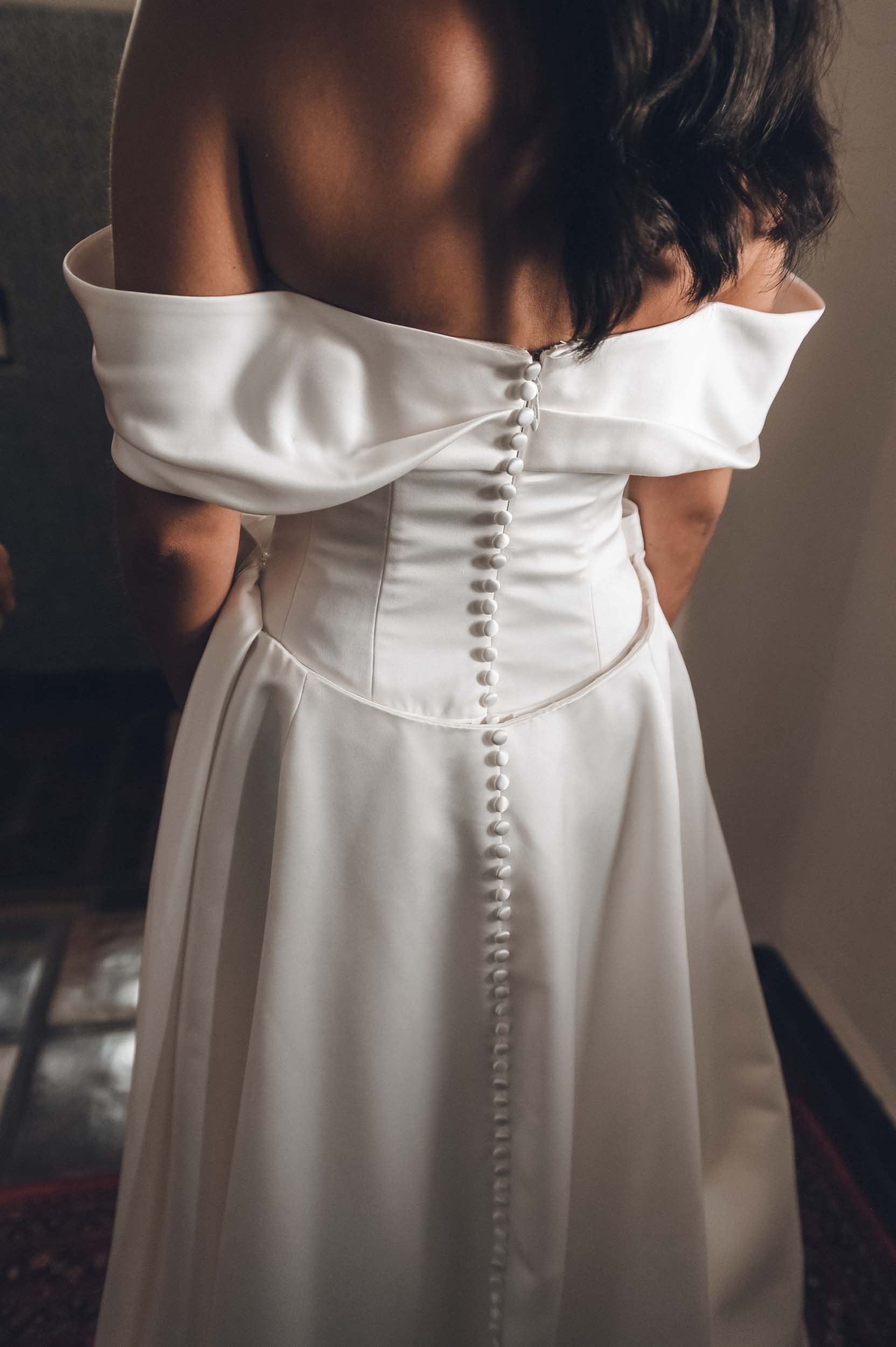 Bride wearing an elegant off-the-shoulder white dress with buttons down the back, standing indoors with dark flooring and blurred background.