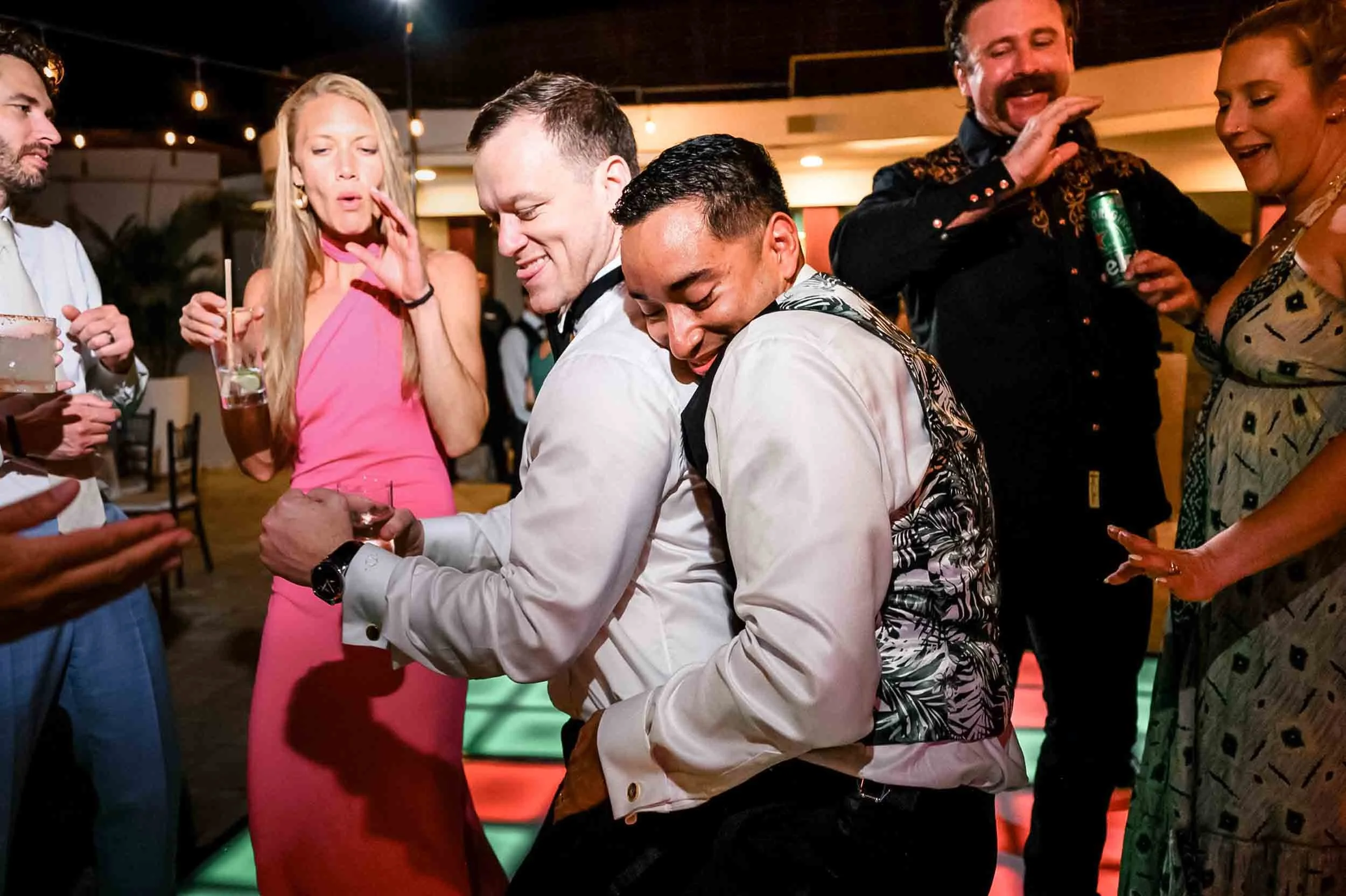 Grooms dancing and socializing at their wedding reception, some are holding drinks, and everyone appears to be enjoying themselves.