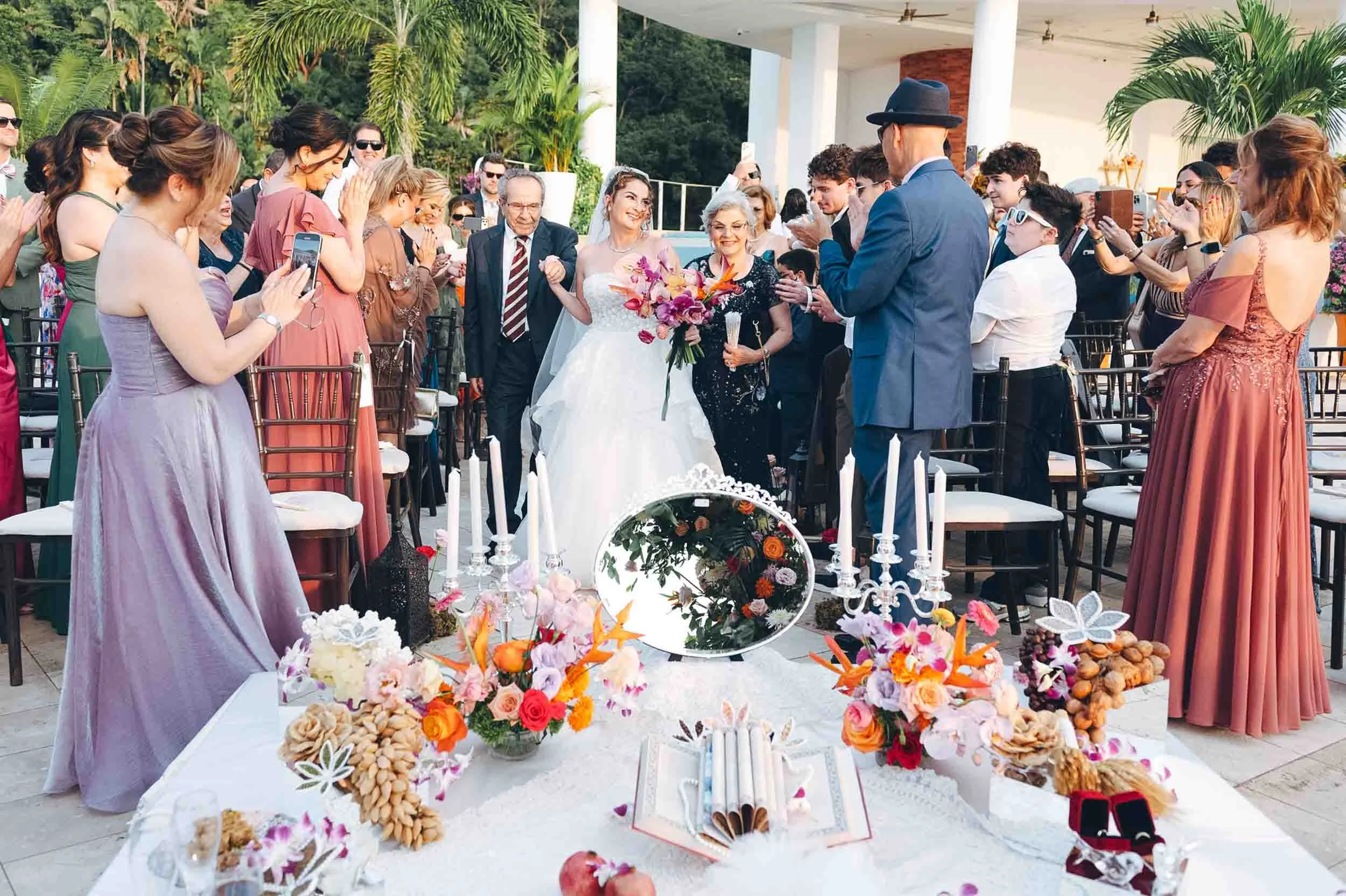 A Persian wedding celebration scene outdoors with a bride in white dress holding a bouquet, walking down the aisle with her parents surrounded by guests clapping and taking photos, and a typical Persian wedding table.
