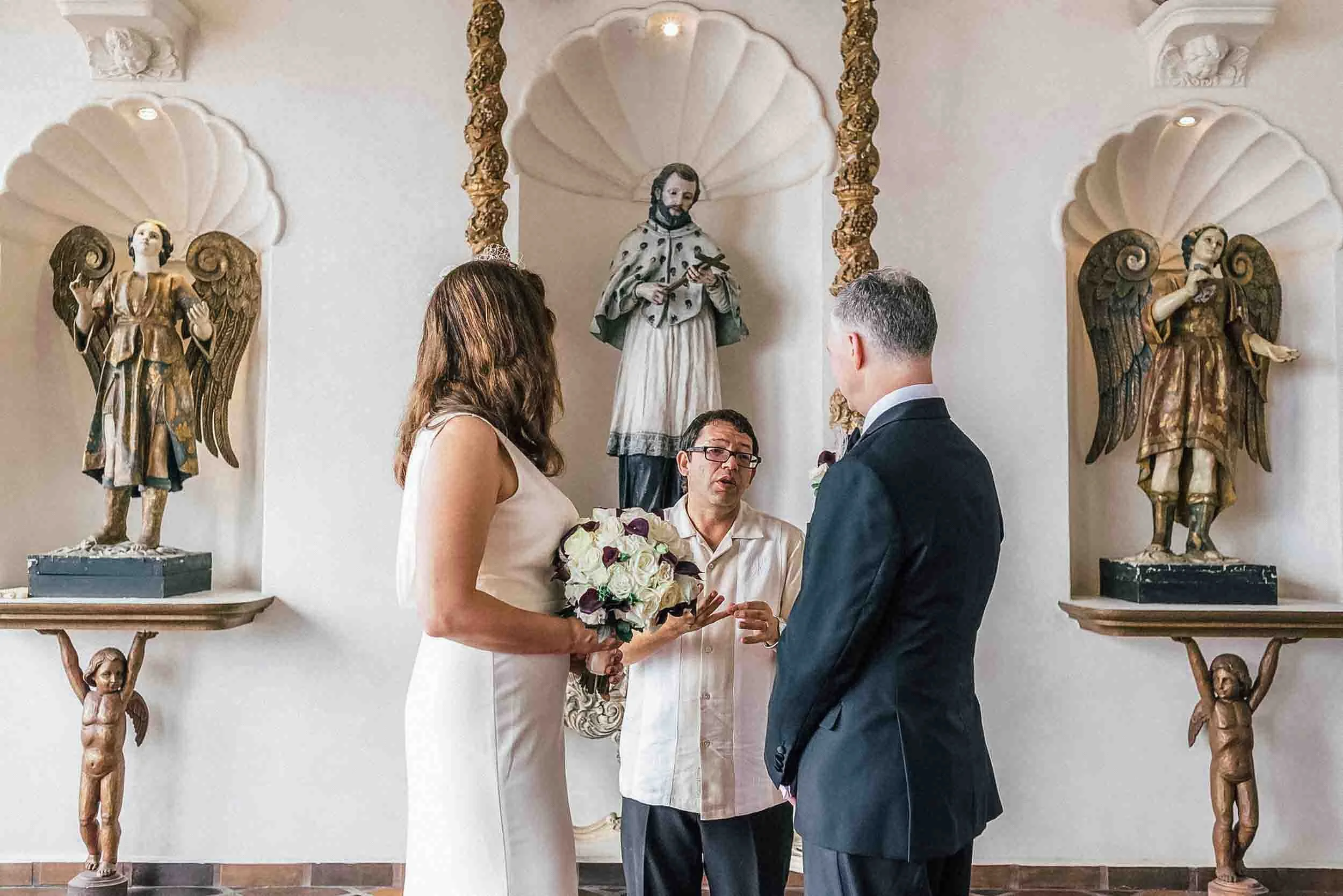 A wedding ceremony taking place  with sculptures of angels on the wall behind the couple and officiant, who is holding a book, while the bride and groom exchange vows.