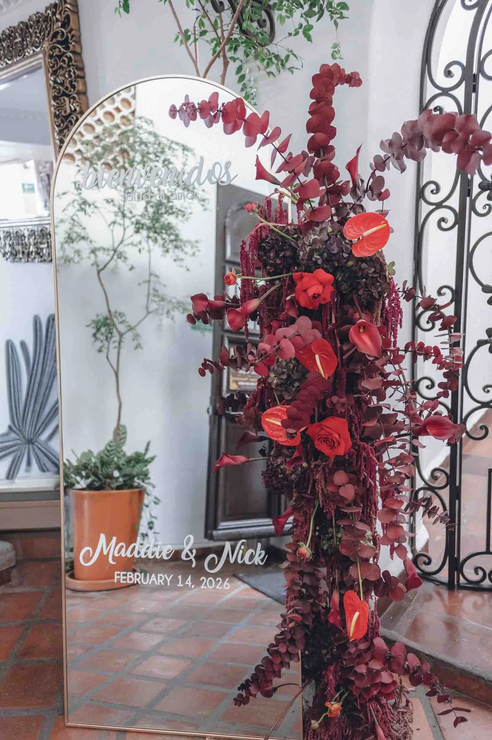 A decorative mirror reflecting a plant with thin branches and small green leaves, with a metal decorative frame in the background. To the right, a tall arrangement of dark red and purple flowers with some orange flowers interspersed, placed in a text