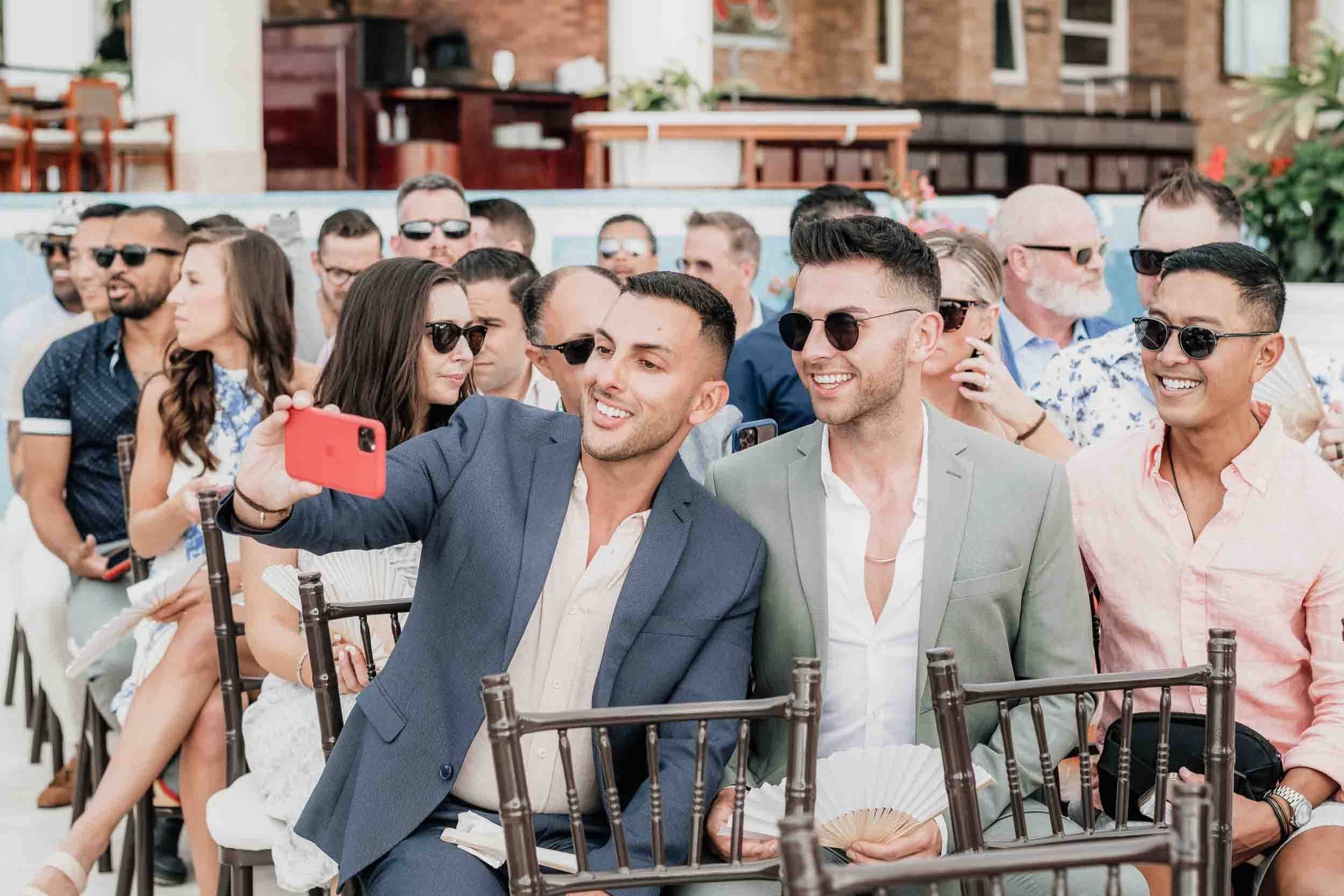 Group of men and women at a gay wedding, taking a selfie with a smartphone. Most are wearing sunglasses, and some are smiling and engaged in conversation. 