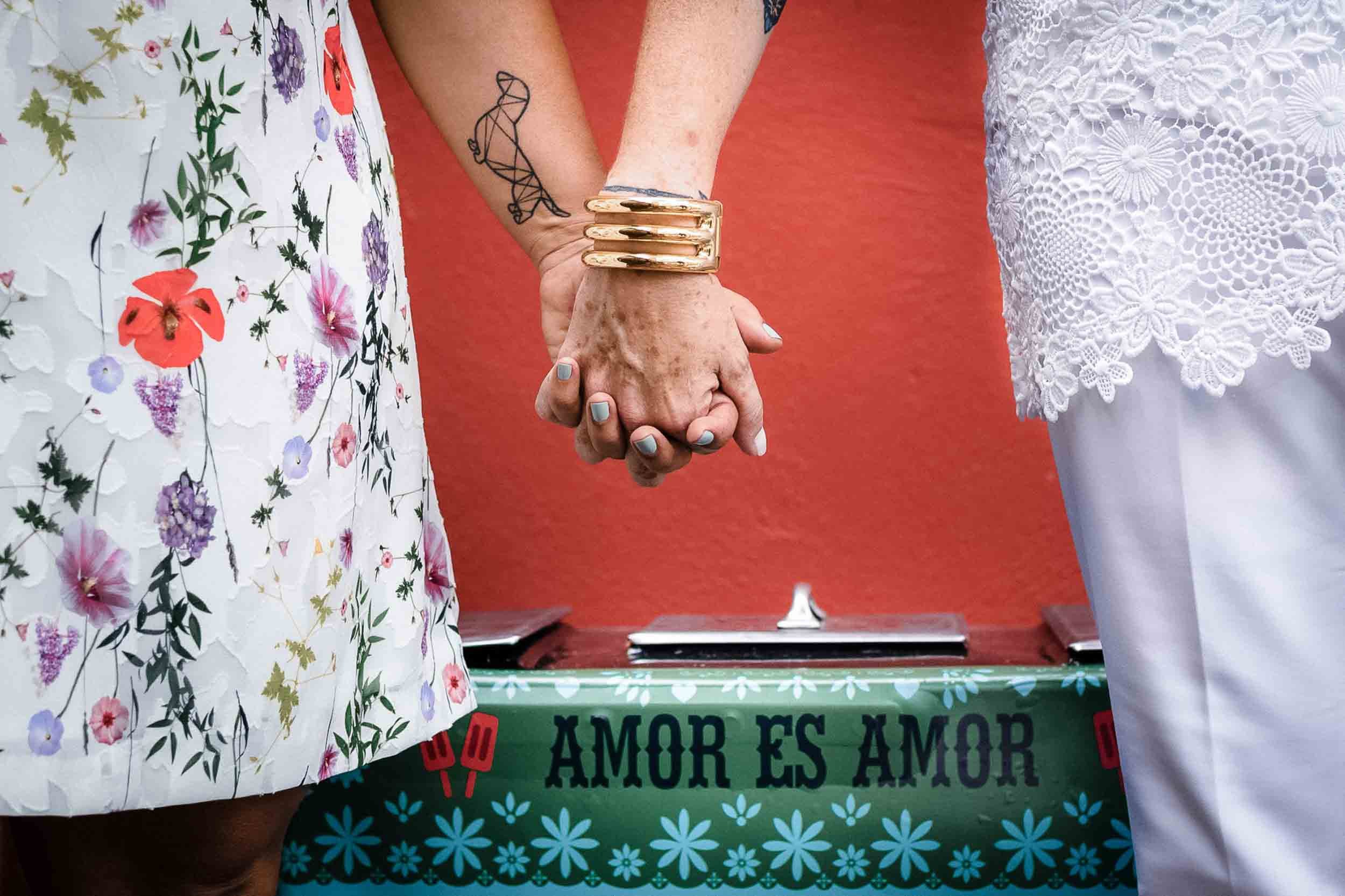Detail of lesbians holding hands with the phrase love is love in the background during  their destination wedding held in Puerto Vallarta Mexico