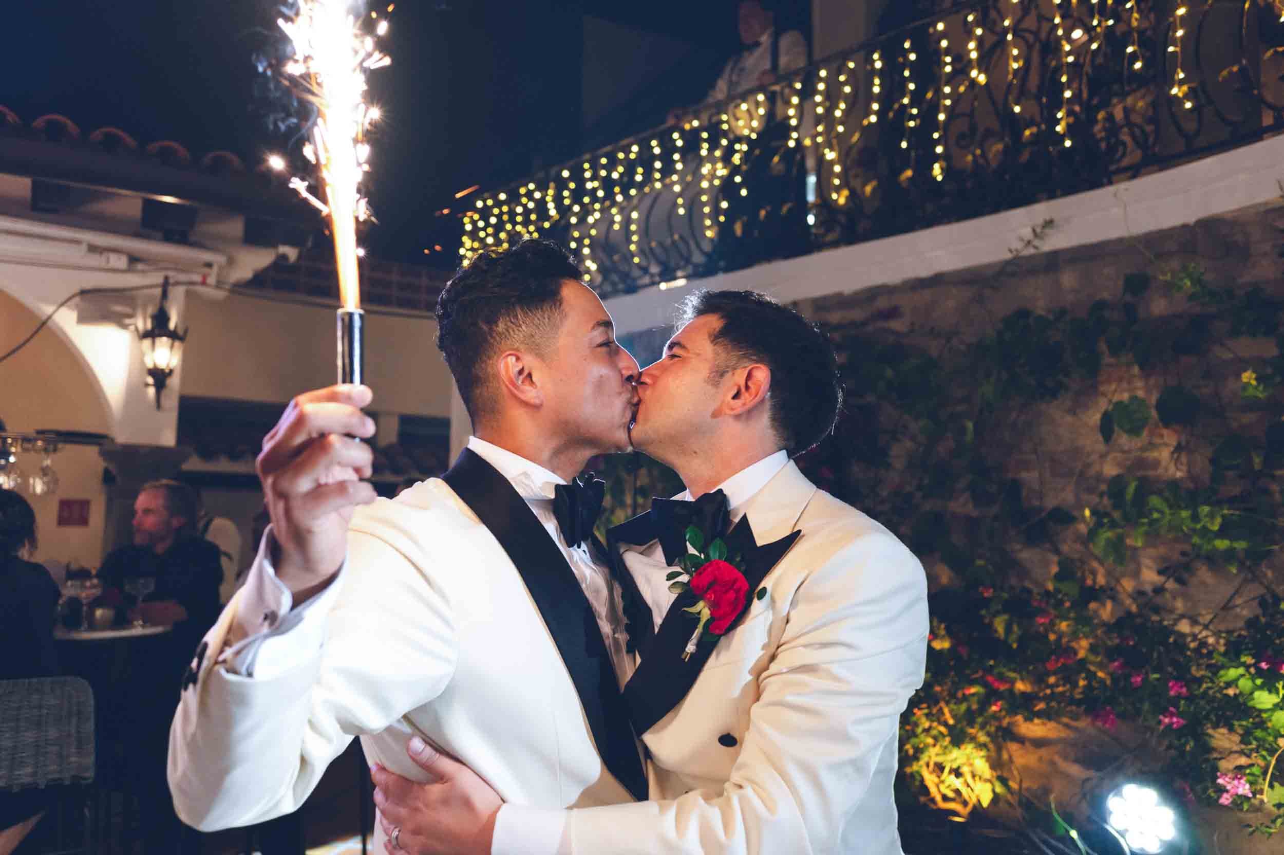 Outdoor setting where gay couple is sharing a kiss at their wedding reception, one holding a sparkler, in an outdoor decorated setting.