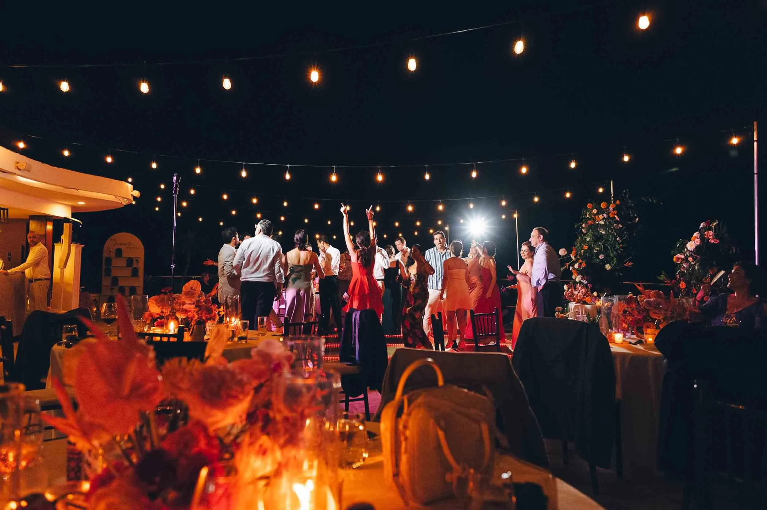 People dancing at an outdoor wedding party decorated with string lights and floral arrangements.