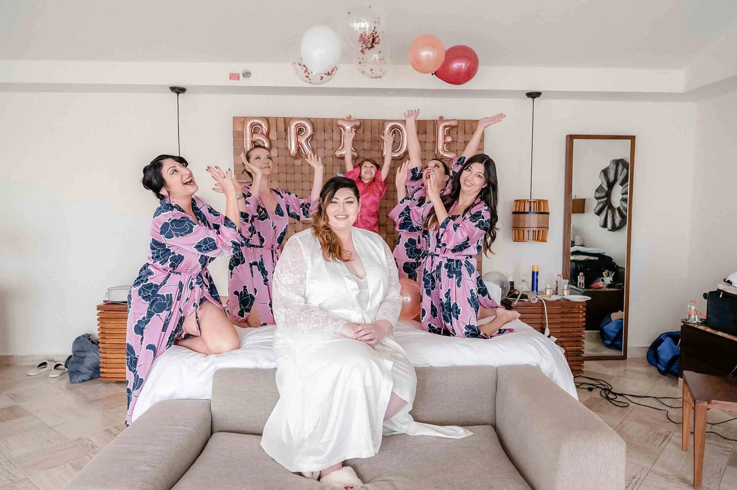 Bride and bridesmaids celebrating in a bedroom with pink and rose gold balloons spelling 'BRIDE' on a wooden headboard, and women wearing matching pink floral robes.