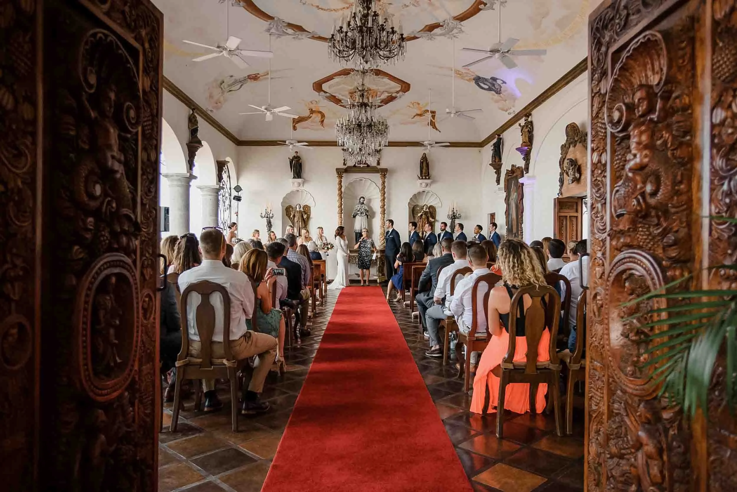 Wedding ceremony at La Capella, surrounded by wooden carvings on both sides, ornate statues, and a ceiling with painted cherubs and a chandelier. 
