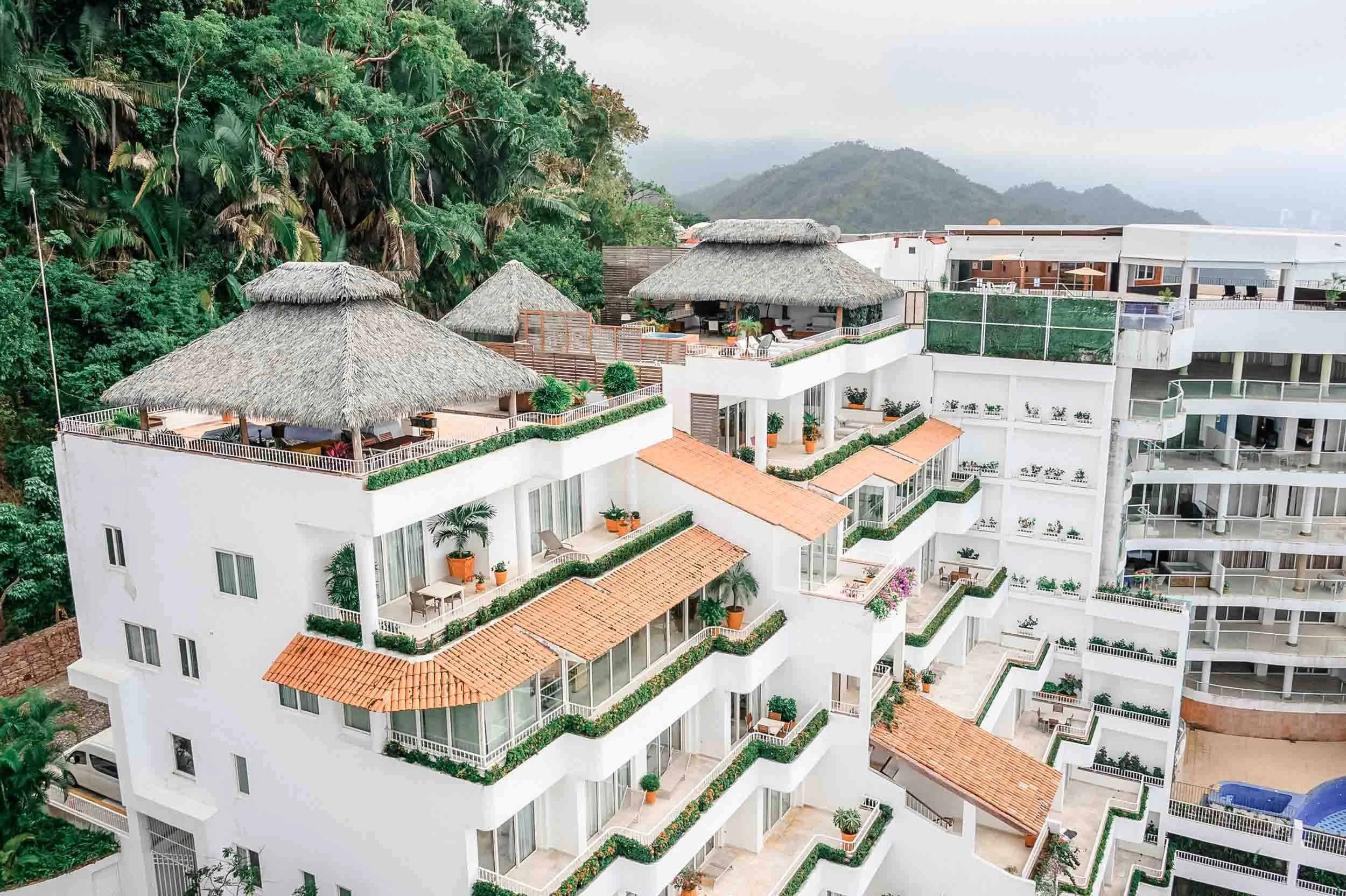Grand Miramar multiple terraces, some with thatched roofs, green plants, and outdoor furniture, surrounded by lush green trees and hills in the background.