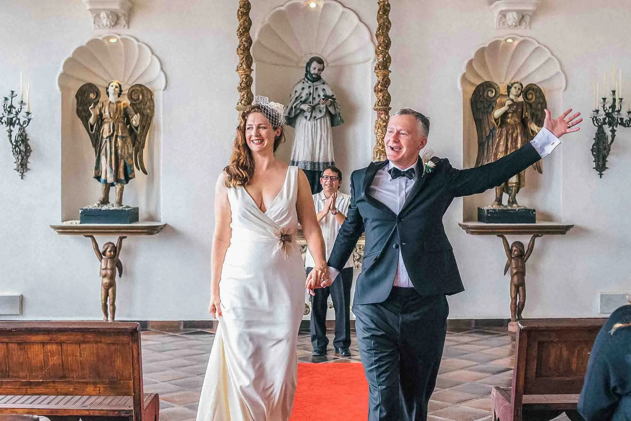 A smiling bride and groom holding hands during their wedding ceremony exit at La Capella. The bride is wearing a white dress with a birdcage veil, and the groom is dressed in a black tuxedo with a bow tie. 