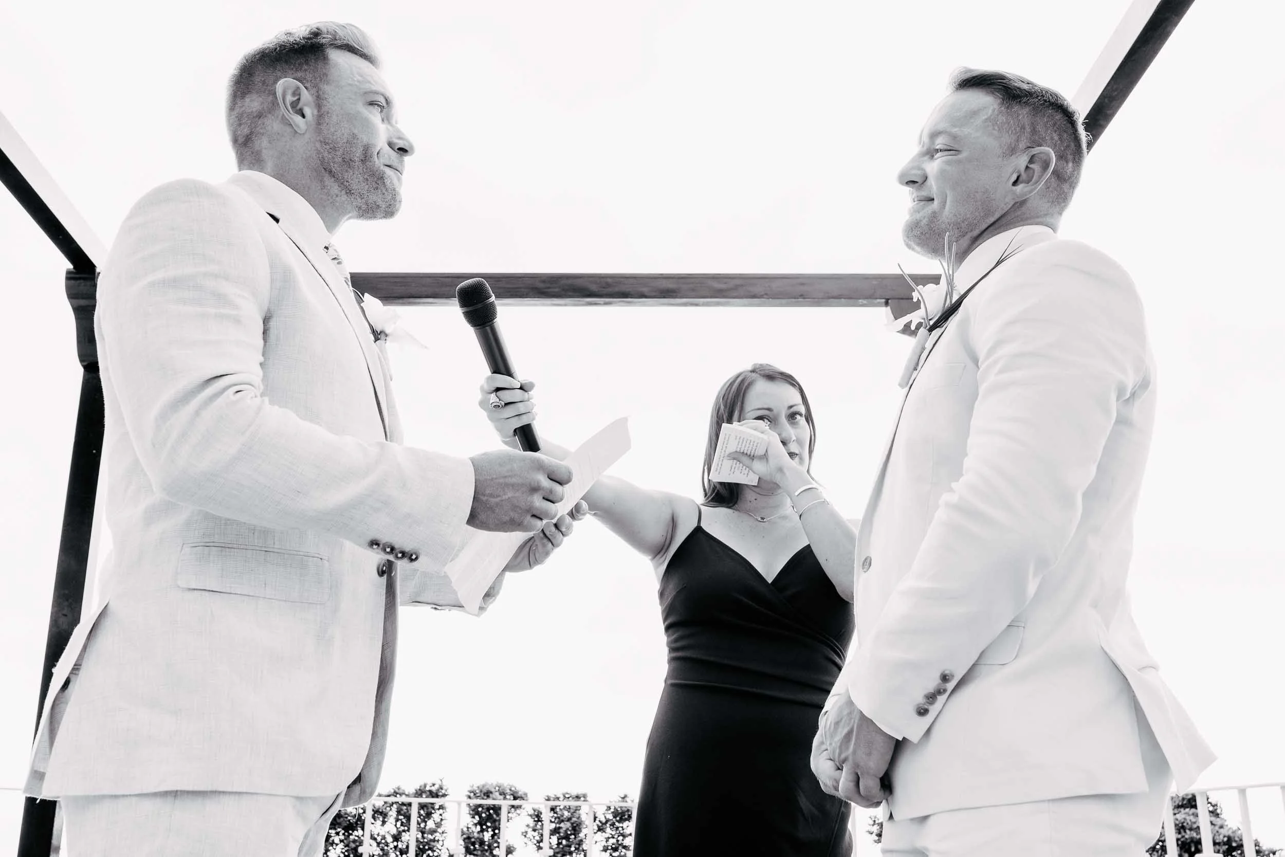 Wedding ceremony with grooms exchanging vows with emotional officiant in the background. 