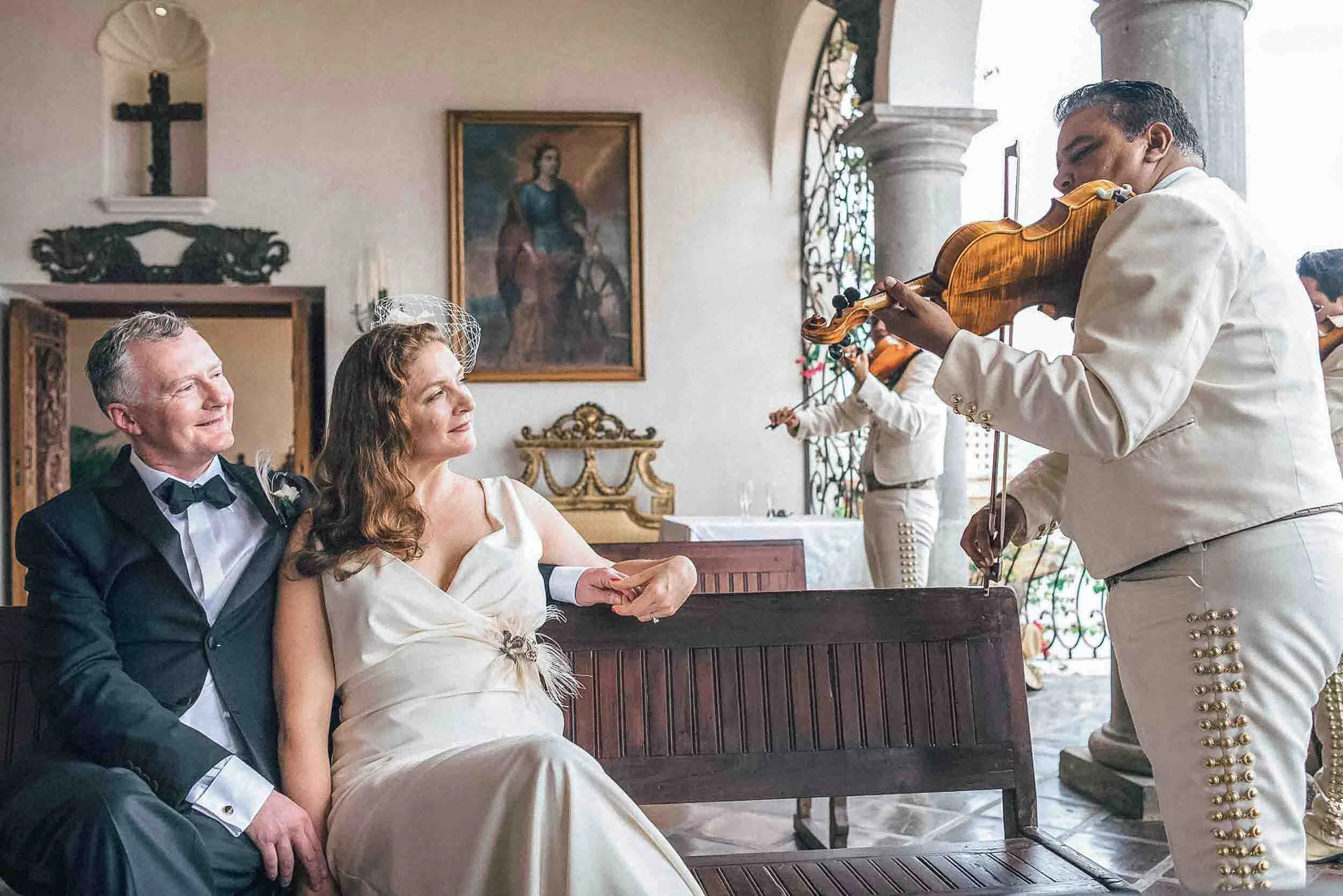 A bride and groom sitting on a pew holding hands enjoying the mariachi music being played for them.